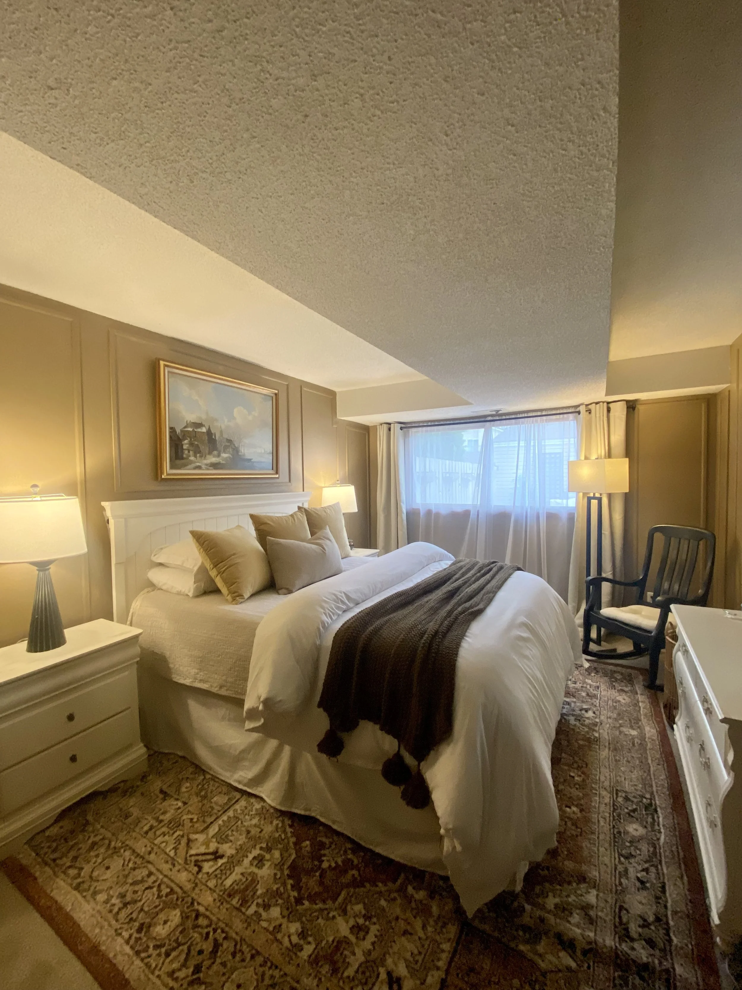 A cozy hotel bedroom with a bed, nightstand, lamps, framed artwork, window with curtains, and a chair, featuring warm lighting and traditional decor.