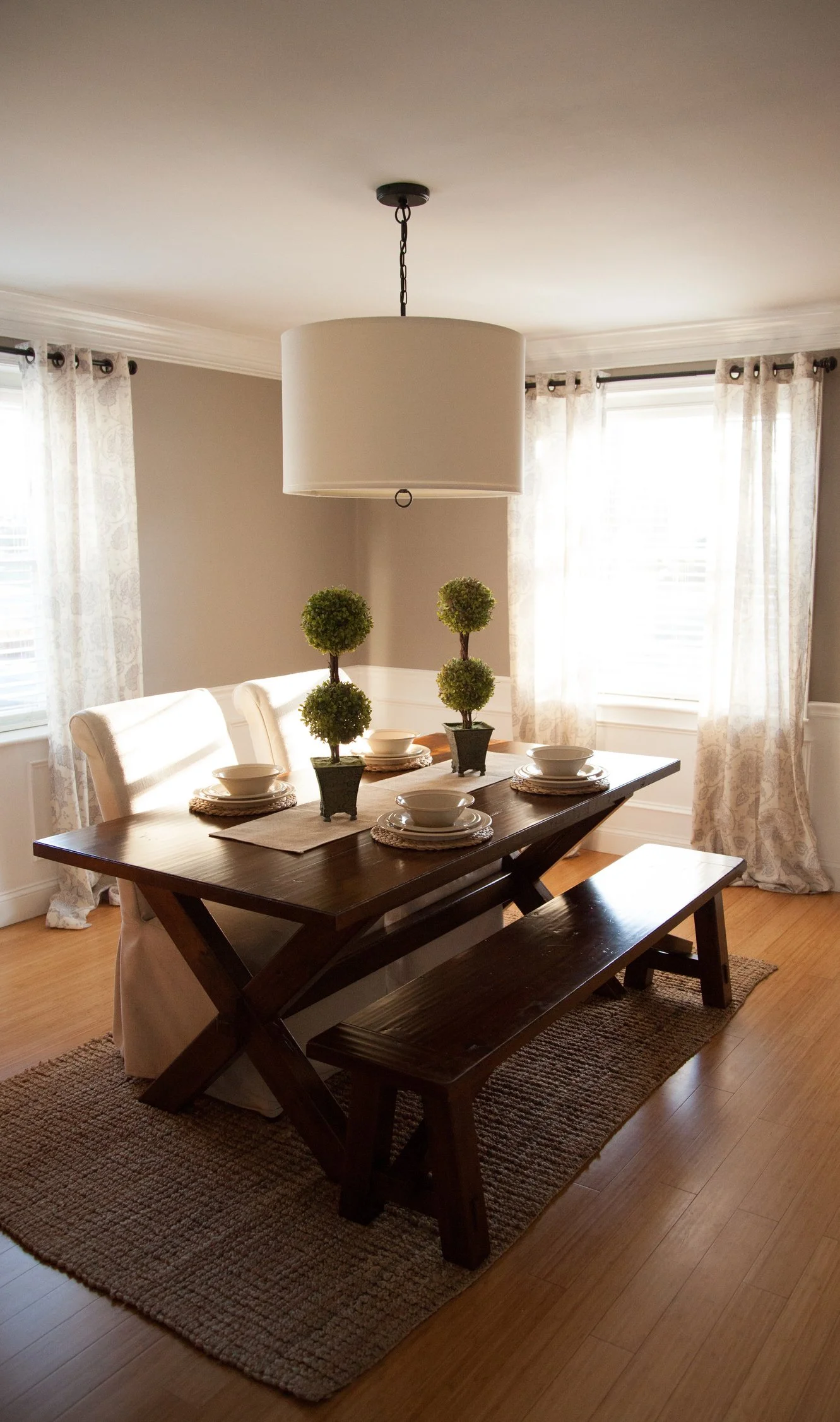 A dining room with a wooden table set with white bowls and plates, topped with a beige table runner, and decorated with three small topiary plants in black pots. There is a white cloth covering the wooden benches. The room has two large windows with sheer curtains and a ceiling light fixture hanging from a chain.