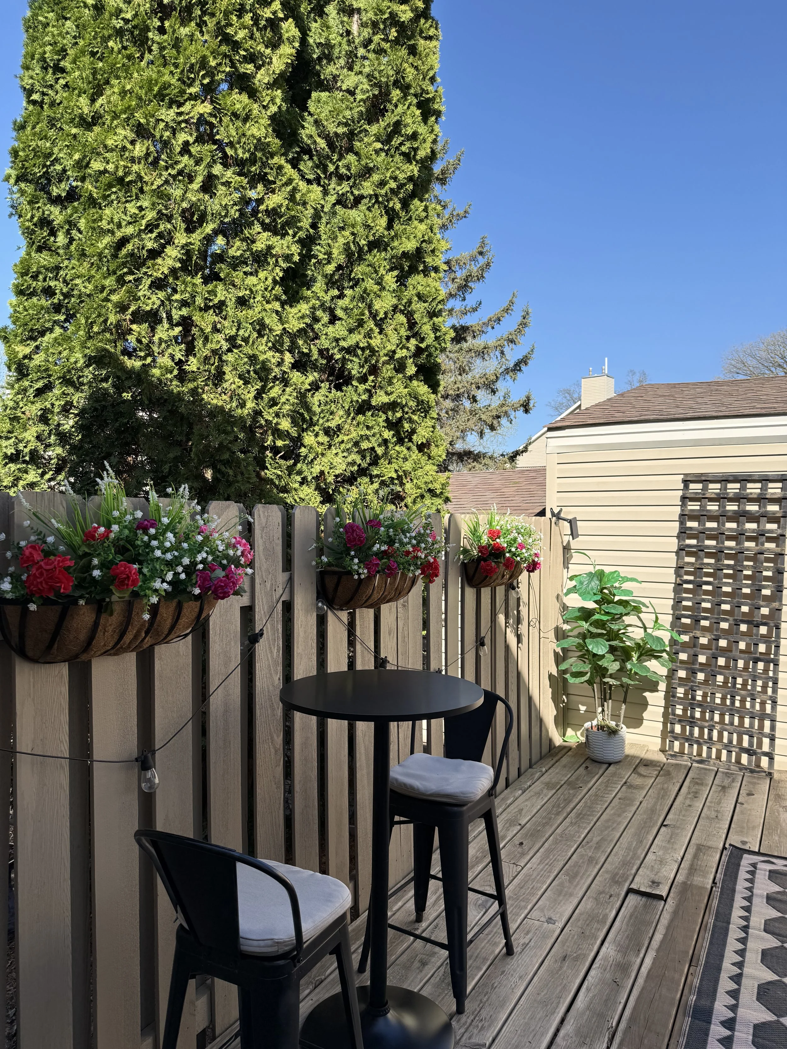 A small outdoor patio with wooden decking, a black table, two black chairs with white cushions, hanging flower baskets with pink and red flowers, a potted plant in a white pot, and a wooden fence with a lattice section. The background shows a large evergreen tree, a house with a pitched roof, and a clear blue sky.
