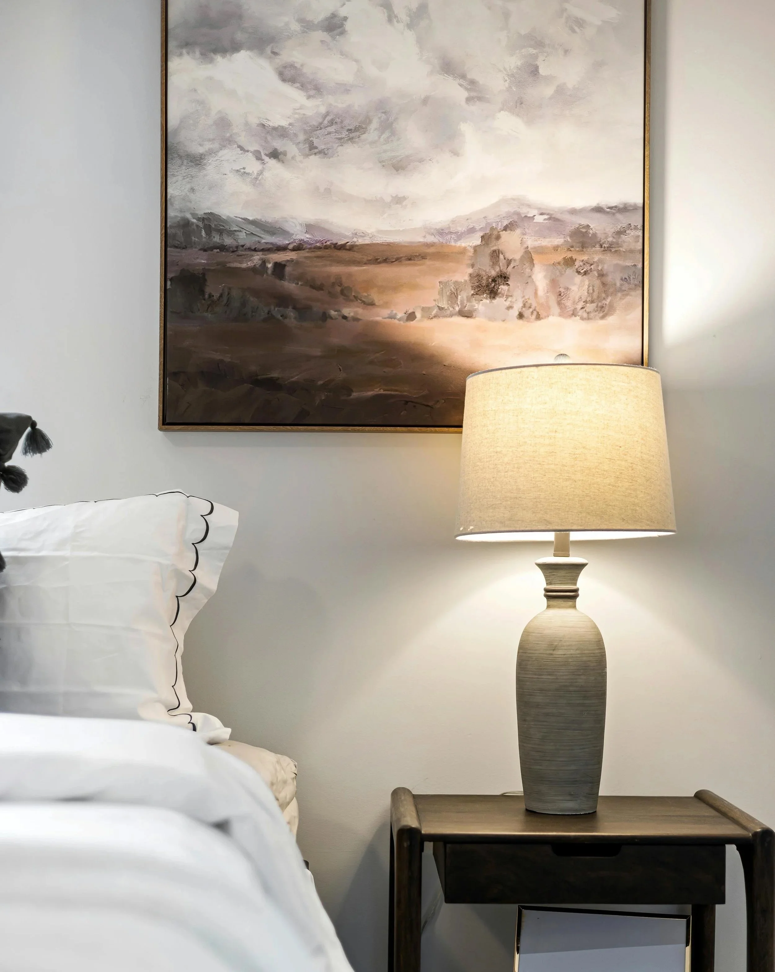 A bedroom with white bedding and pillows on the left, a bedside table with a large gray textured vase, a beige lampshade, a cream-colored lamp, and a landscape painting with mountains and clouds on the wall.
