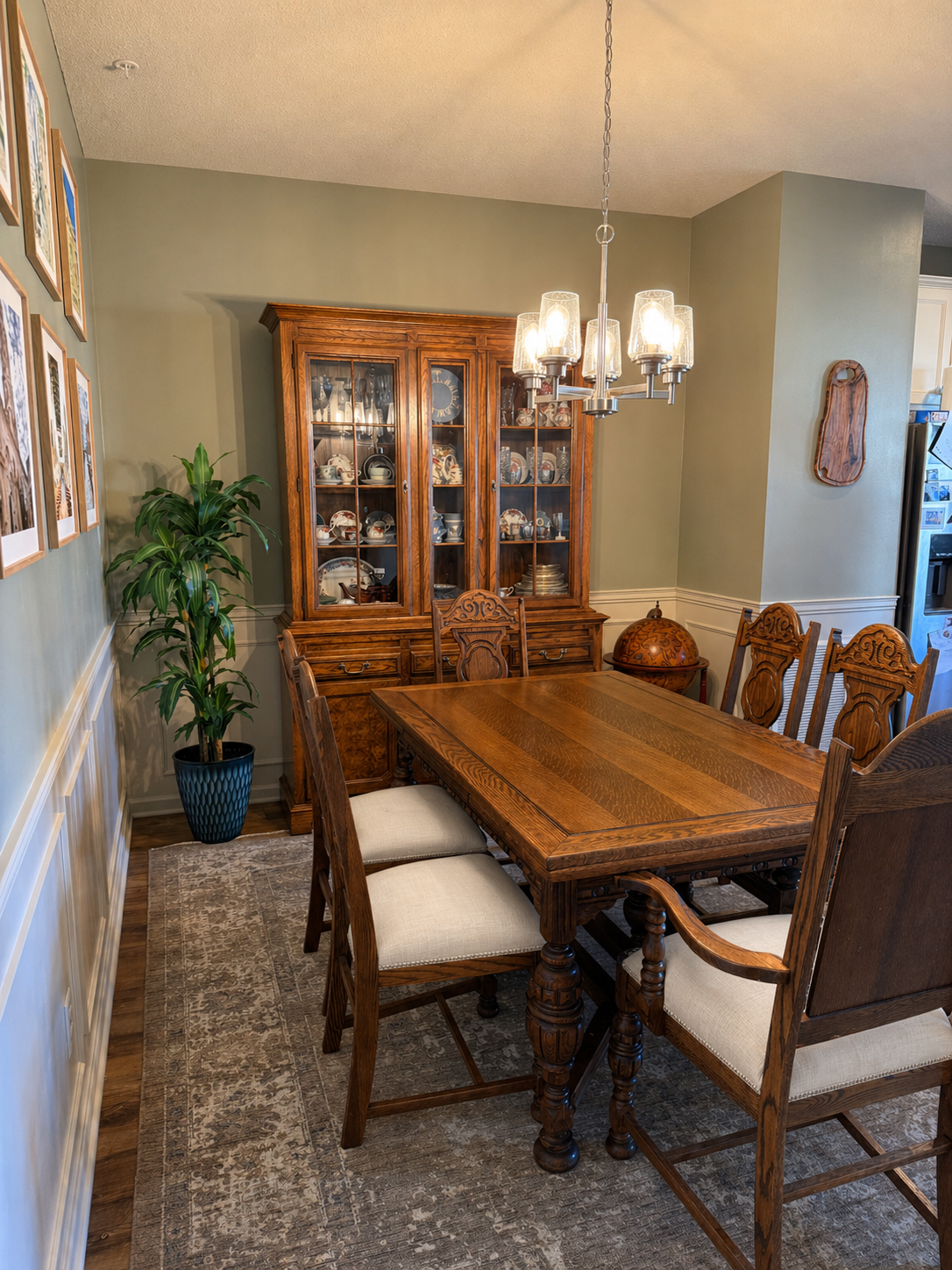 Dining room with wooden table, six matching chairs with beige cushions, a large wooden china cabinet with glass doors displaying dishes, a green potted plant in a blue patterned pot, a vintage globe on a stand, a chandelier above the table, and framed pictures on the wall.