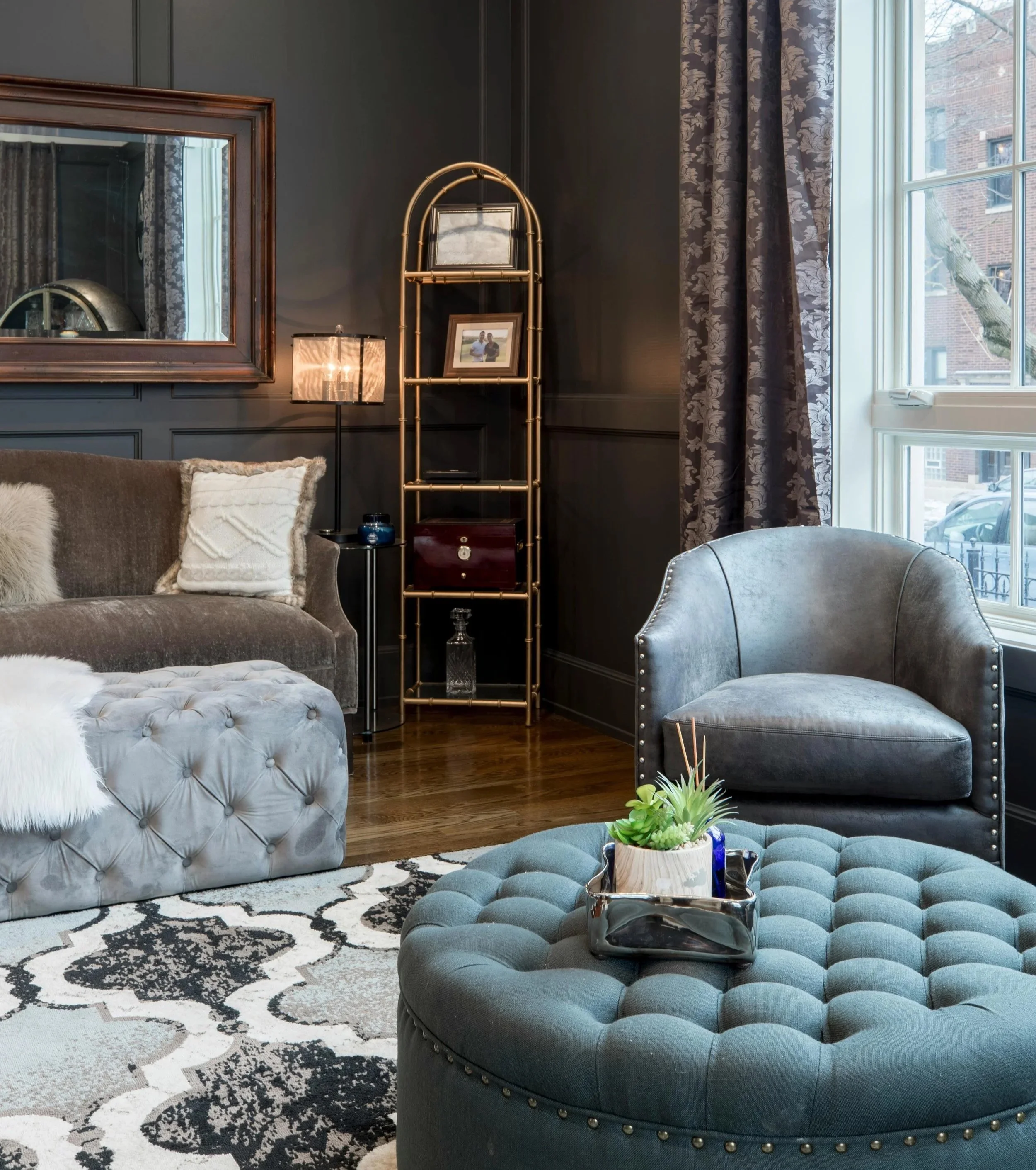 A cozy living room corner with a gray tufted ottoman, a gray upholstered armchair, a dark patterned rug, a black wall, a large window with floral curtains, a gold shelving unit with framed photos, a table lamp, and a small potted plant on a tray.