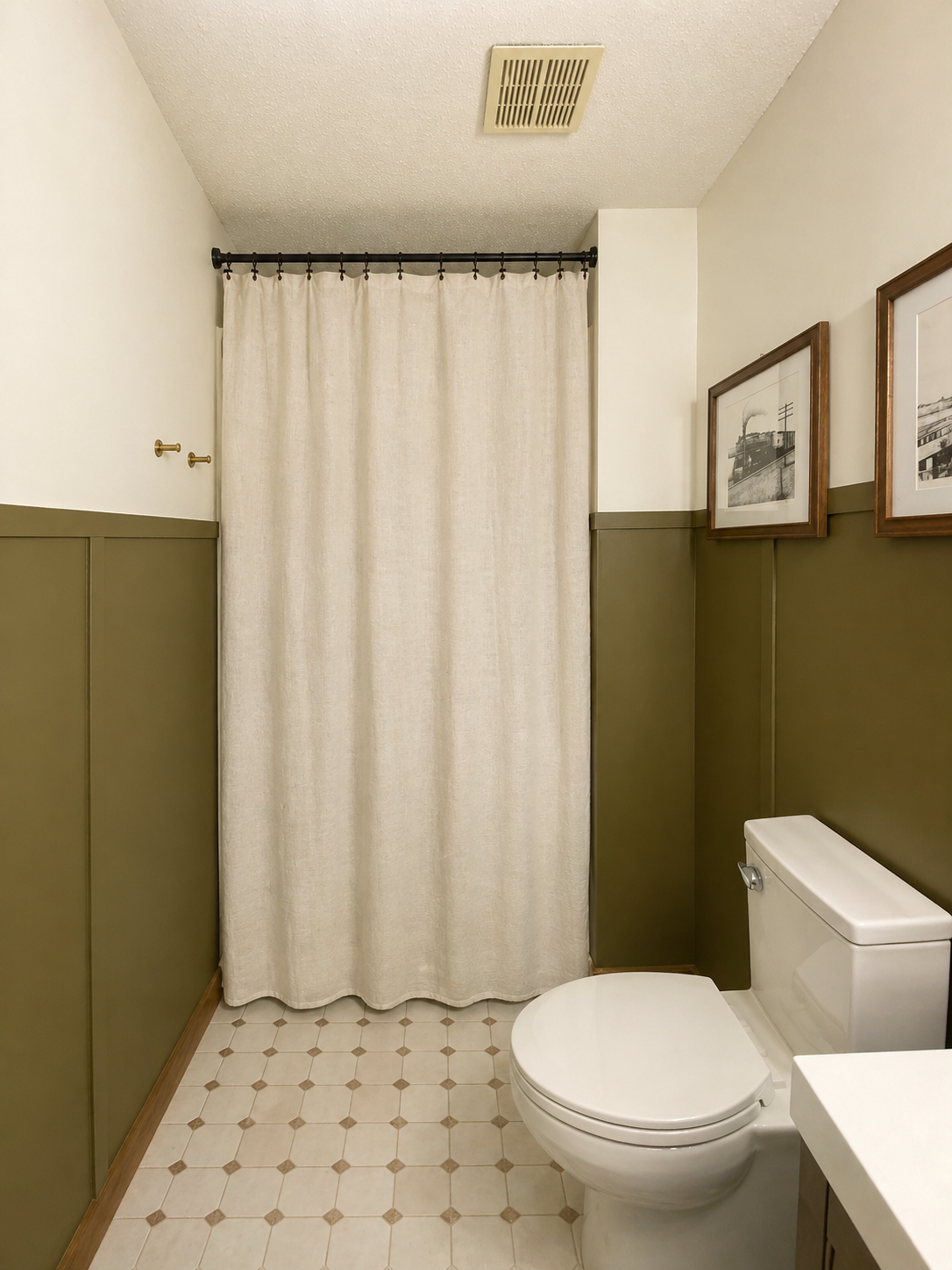 A small bathroom with a toilet, a beige shower curtain, framed black-and-white photos on the wall, and green wainscoting on the walls.