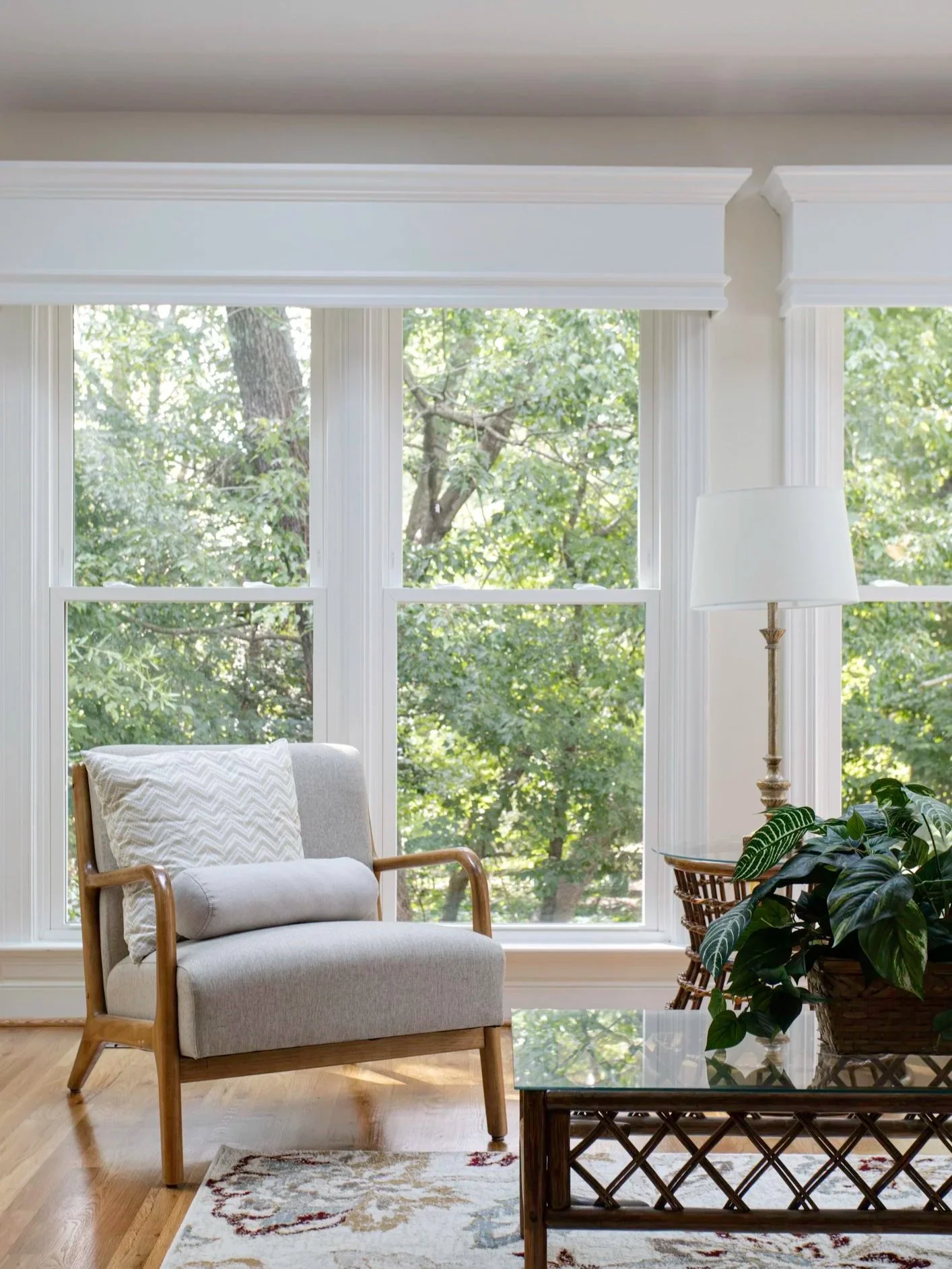 Living room with large window showing green trees outside, beige armchair with wooden arms and a chevron-patterned pillow, glass-topped coffee table with woven base, standing white floor lamp, and potted plant, with a patterned rug on hardwood floor.