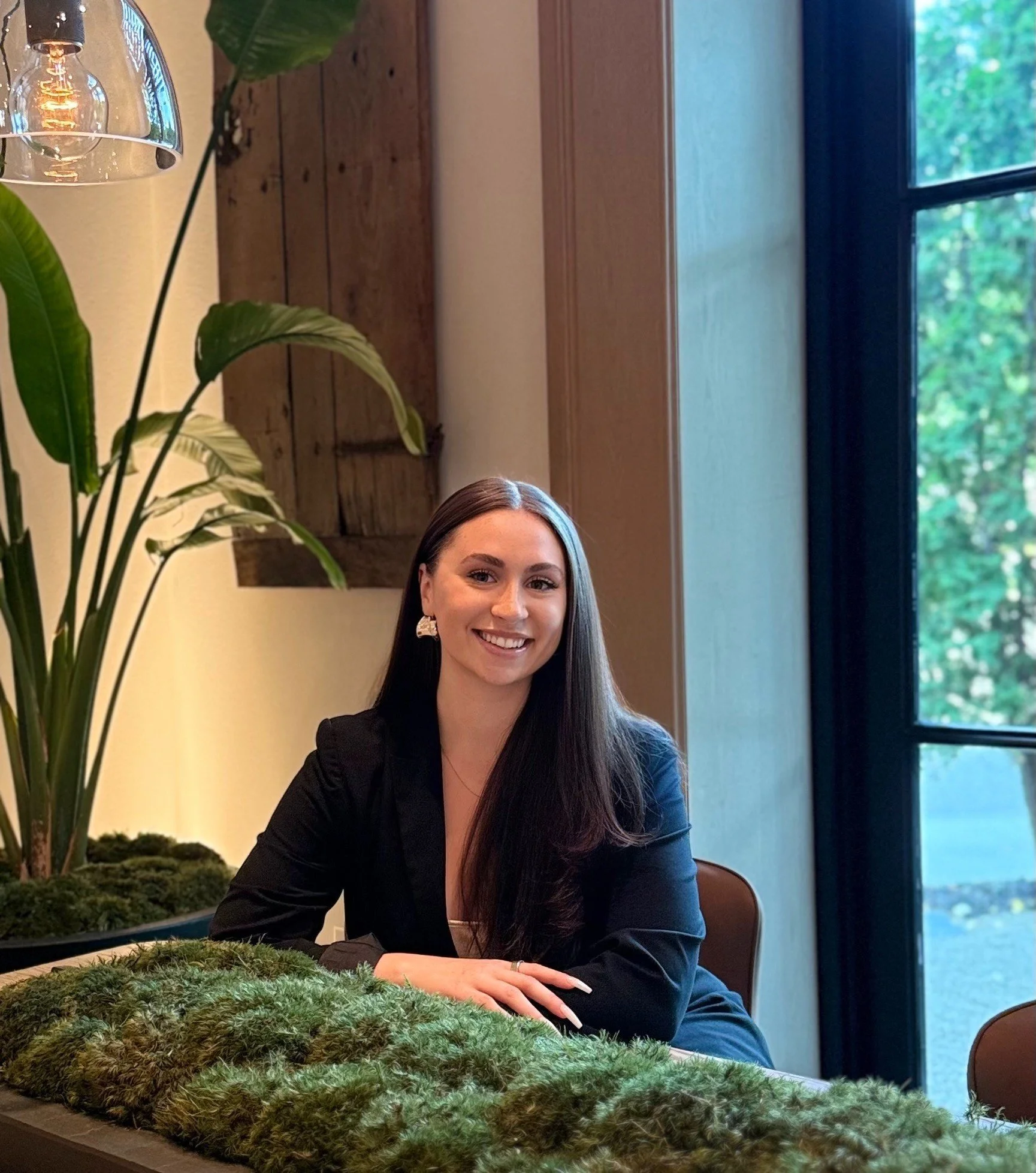A woman with long dark hair, smiling and wearing a black blazer, sitting at a table in a room with a large window showing trees outside. There is a green leafy plant and moss on the table in the foreground, with a wooden wall and a window in the background.