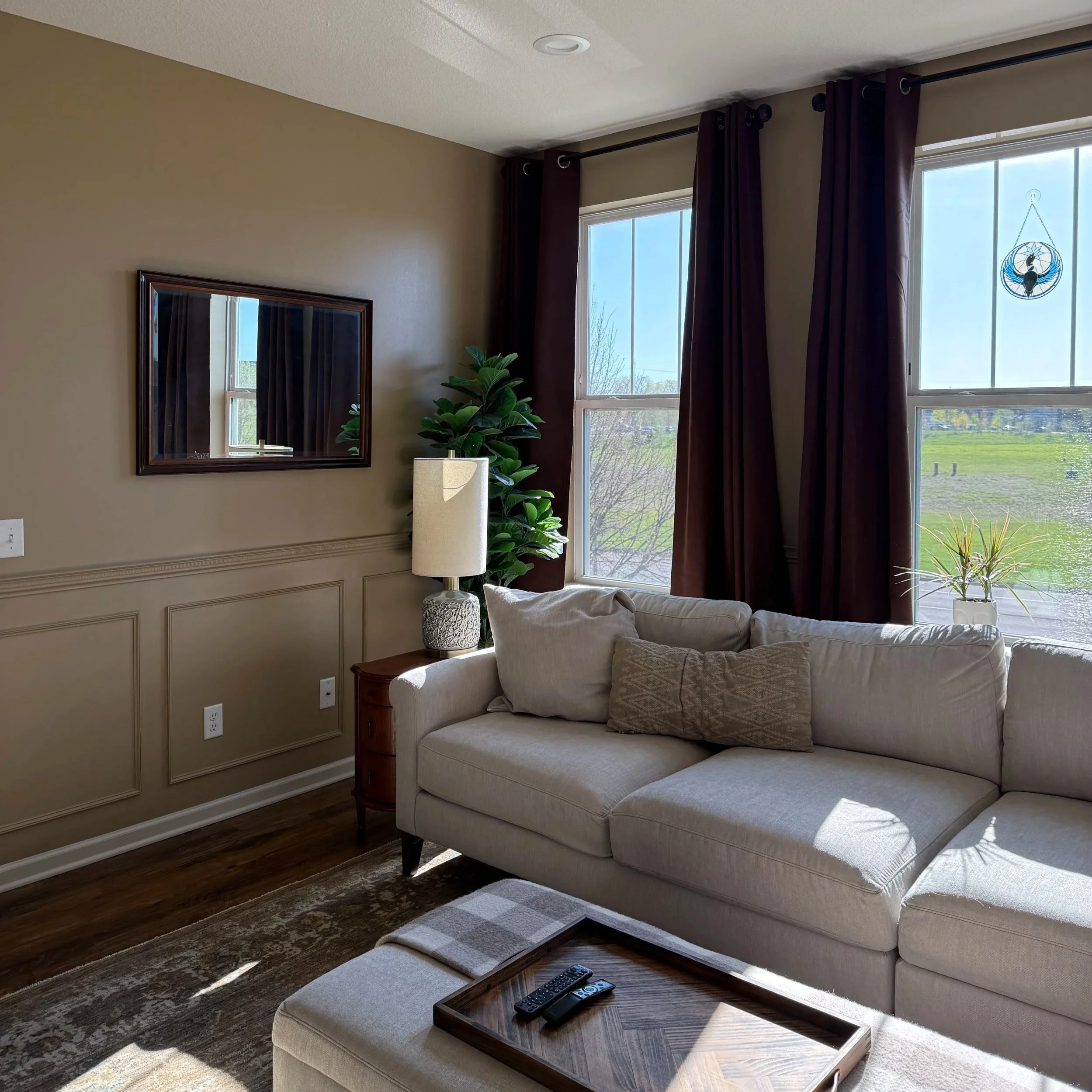 Living room with beige walls, two large windows with dark curtains, a beige sofa with a decorative pillow, a wooden coffee table with remote controls, a lamp, a green potted plant, a wooden side table, a rug, a wall mirror, and a decorative hanging piece.
