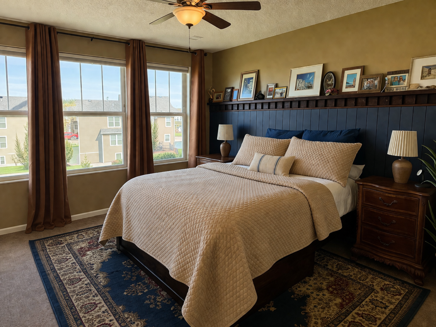 A cozy bedroom with a large bed, beige quilt, and multiple pillows, flanked by nightstands with lamps. There are three windows with brown curtains, a ceiling fan, and a dark blue wall with framed artwork above the bed.