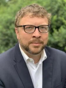 Adventure Scholars advisor Dr. Robby Luckett wearing glasses and a suit outdoors with greenery in the background.