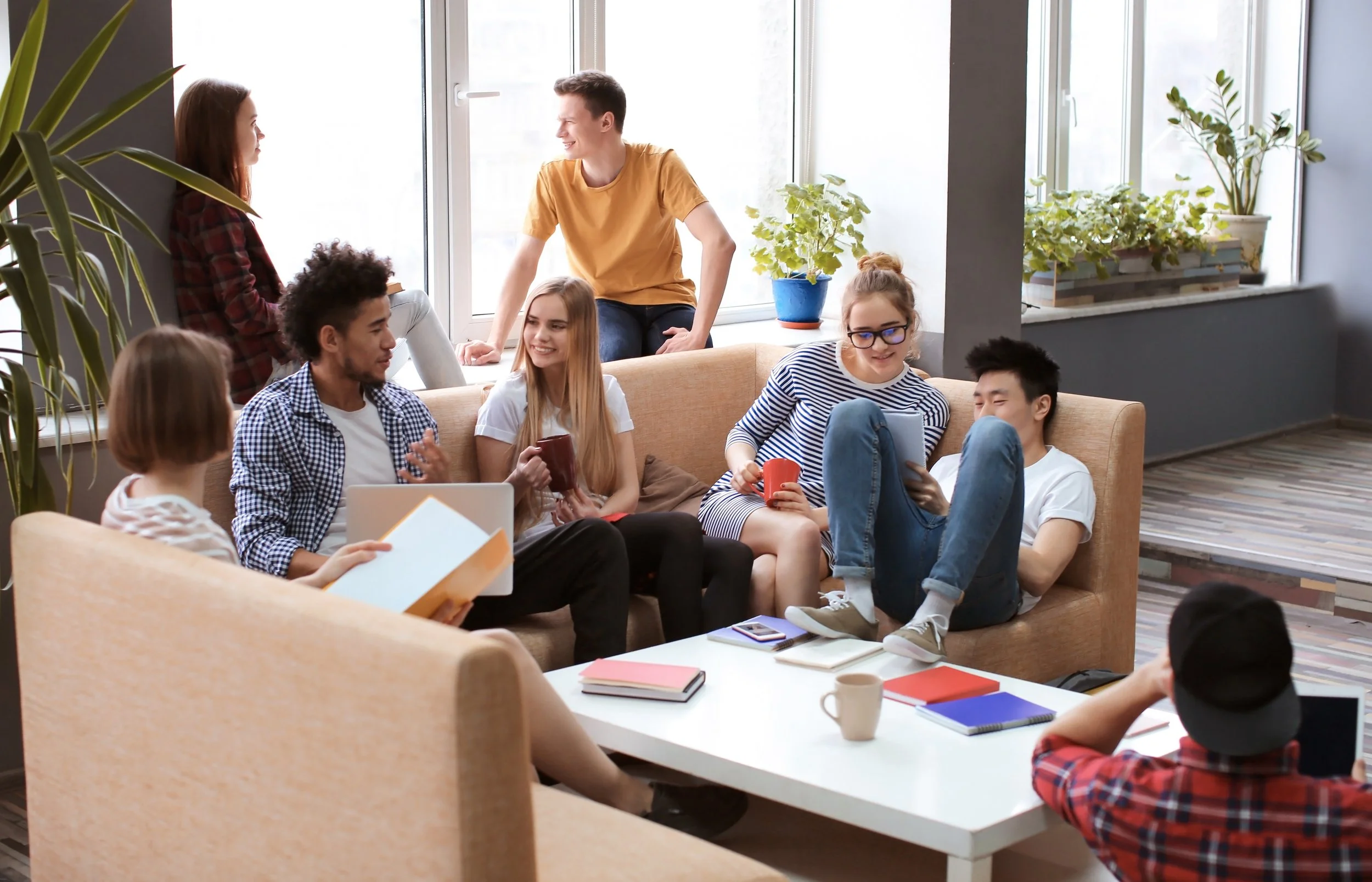 Group of young adults socializing in a modern lounge area, sitting on sofas with laptops and books, discussing what they've learned on the tour.