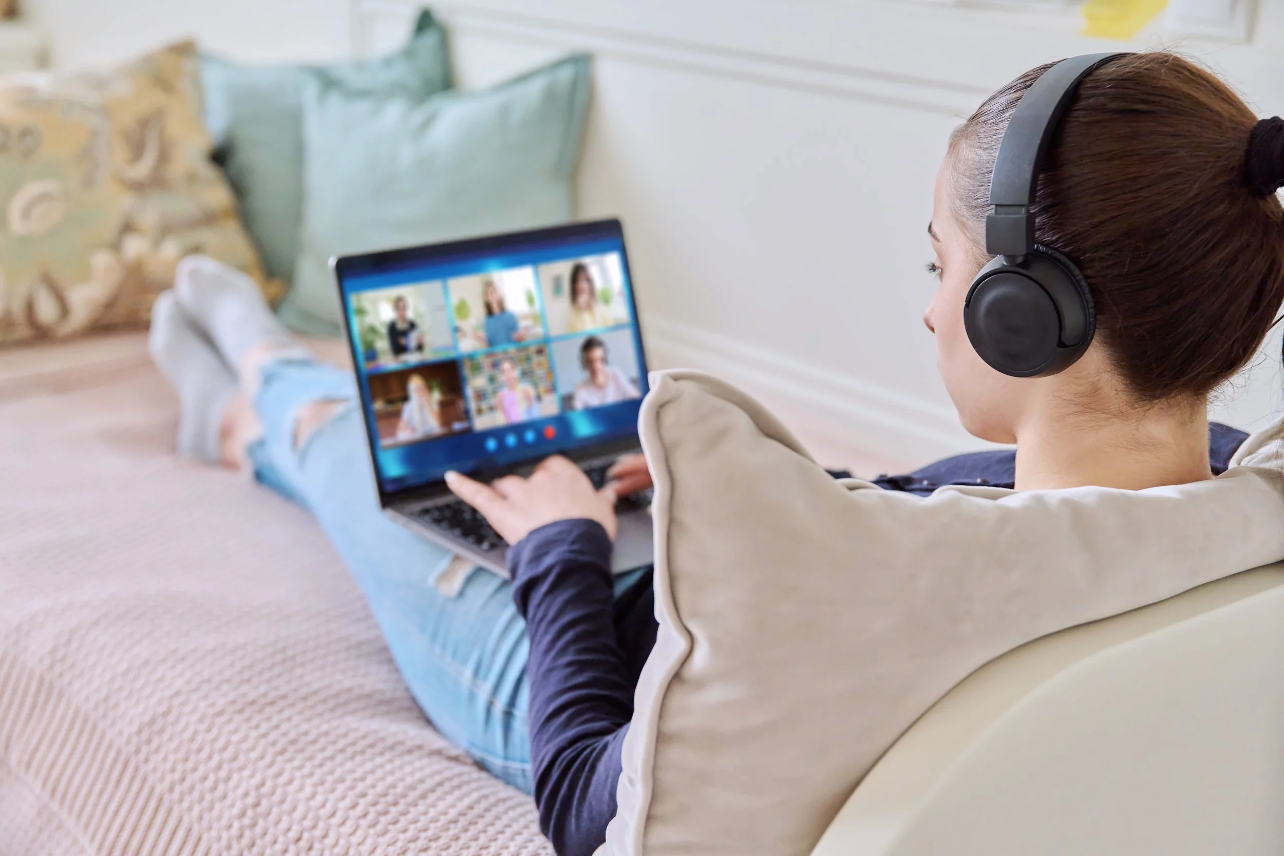 A student wearing headphones participating in a video call on a laptop, lying on a bed with pillows.