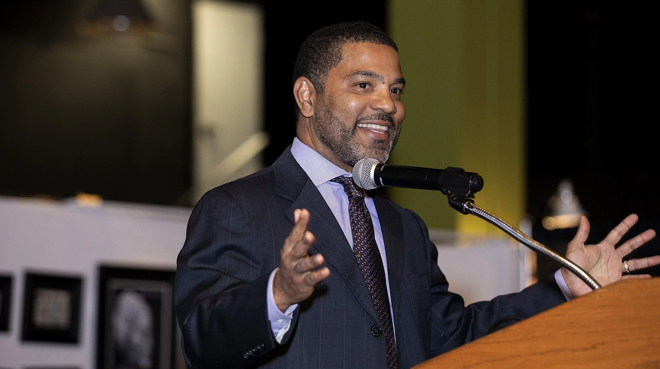 Adventure Scholars co-founder, Robin Perry giving a speech at a podium, wearing a suit and tie, speaking into a microphone.