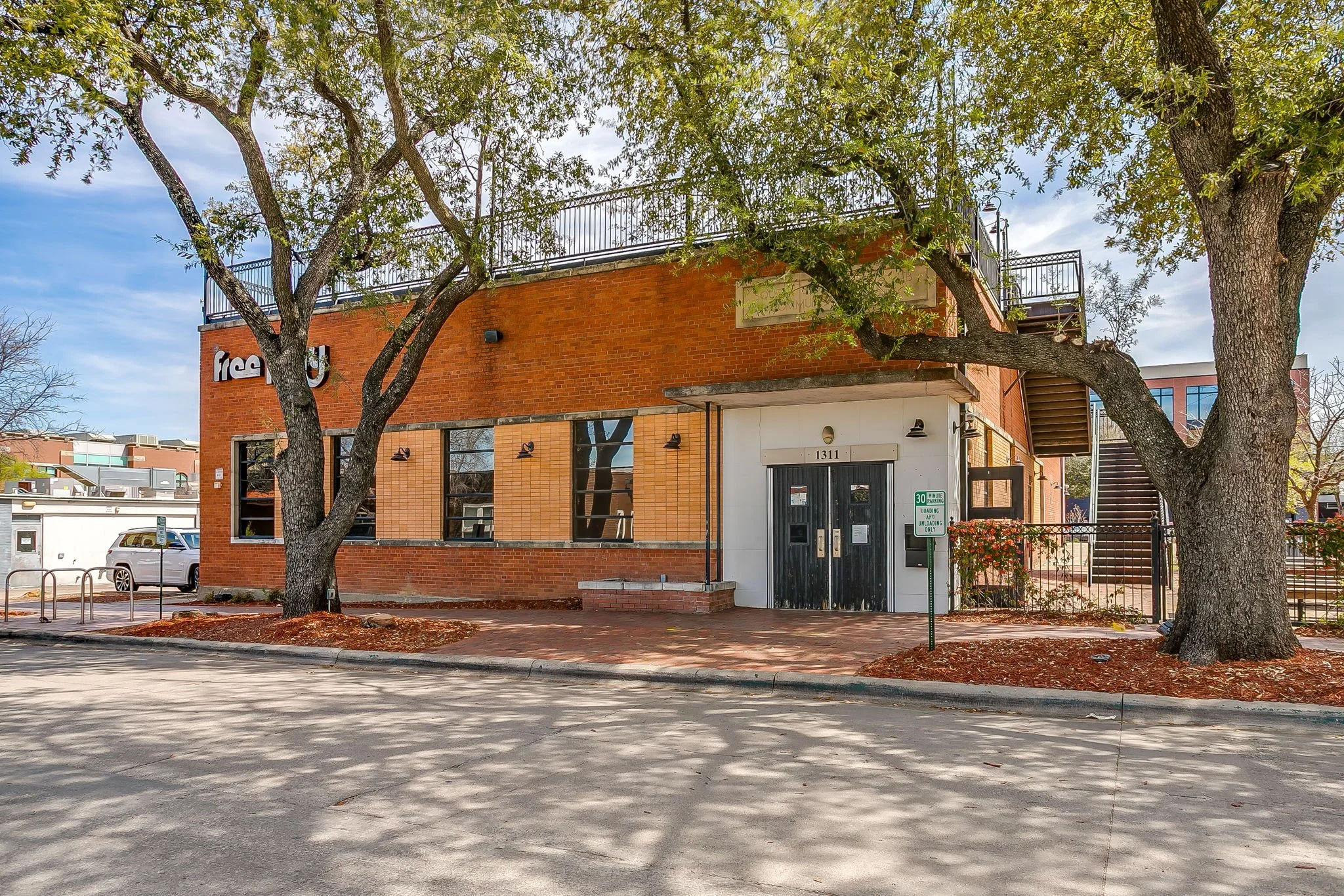 A two-story brick building with black doors and windows, surrounded by large trees with green leaves, situated on a street with parked cars and a bike rack, under a partly cloudy sky.