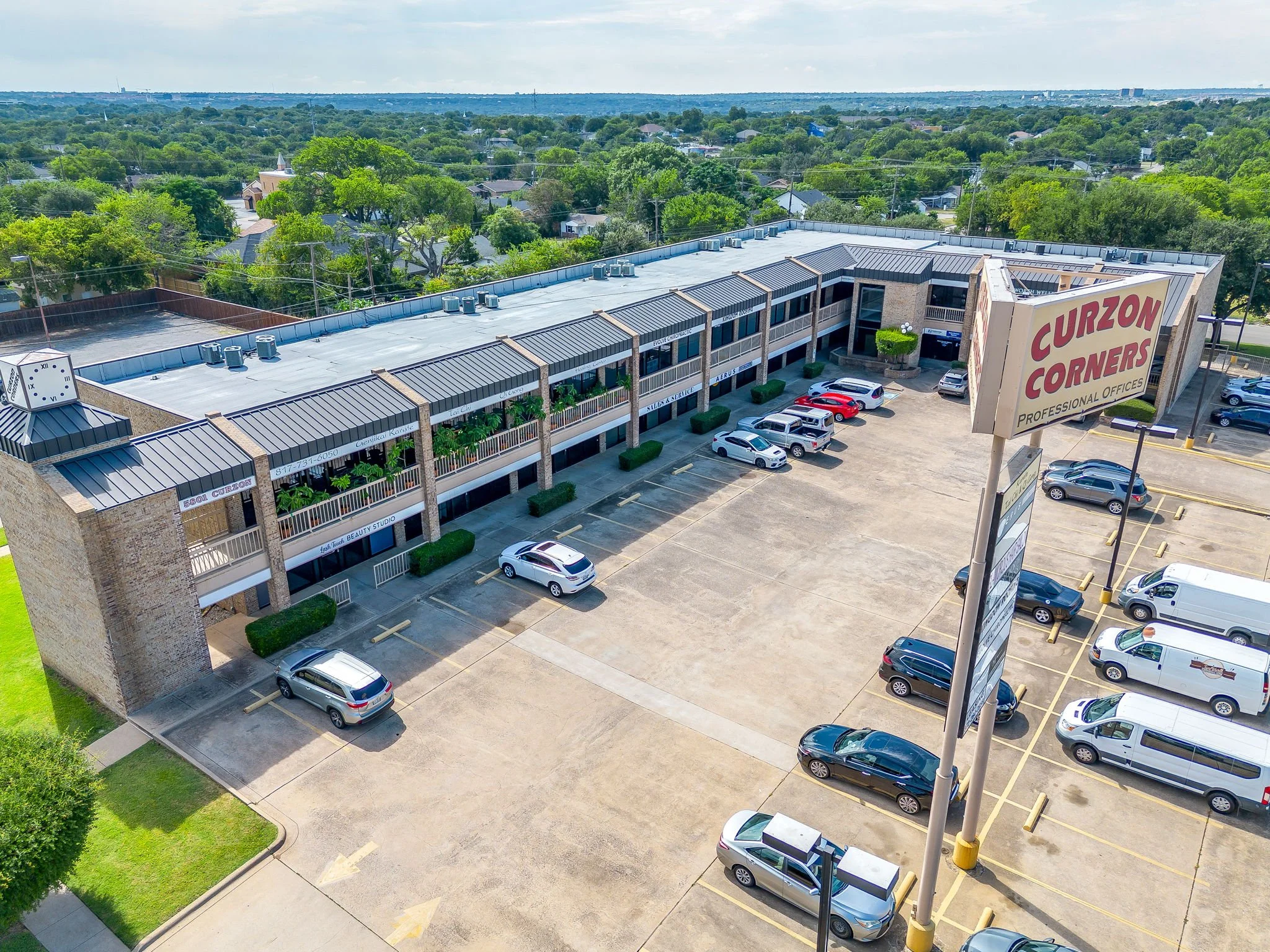 An aerial view of a commercial shopping center parking lot with several parked cars, adjacent to a building labeled "Curzon Corners Professional Offices". The background shows a residential neighborhood with lush green trees and distant cityscape.