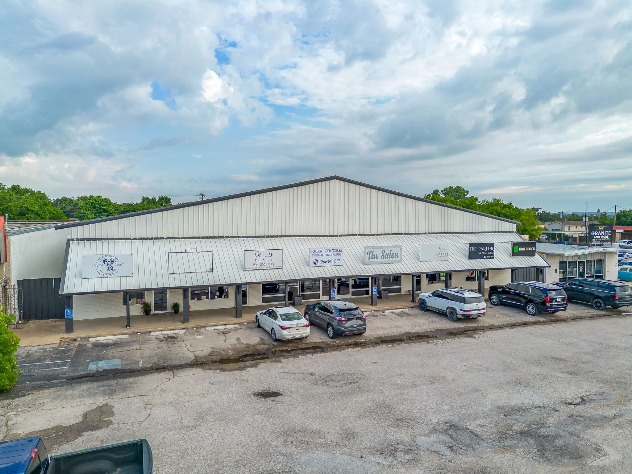A strip mall with a row of small businesses on a cloudy day, including a hair studio, therapeutic massage, salon, and retail stores. Cars are parked in front of the stores, and a parking lot is visible in the foreground.