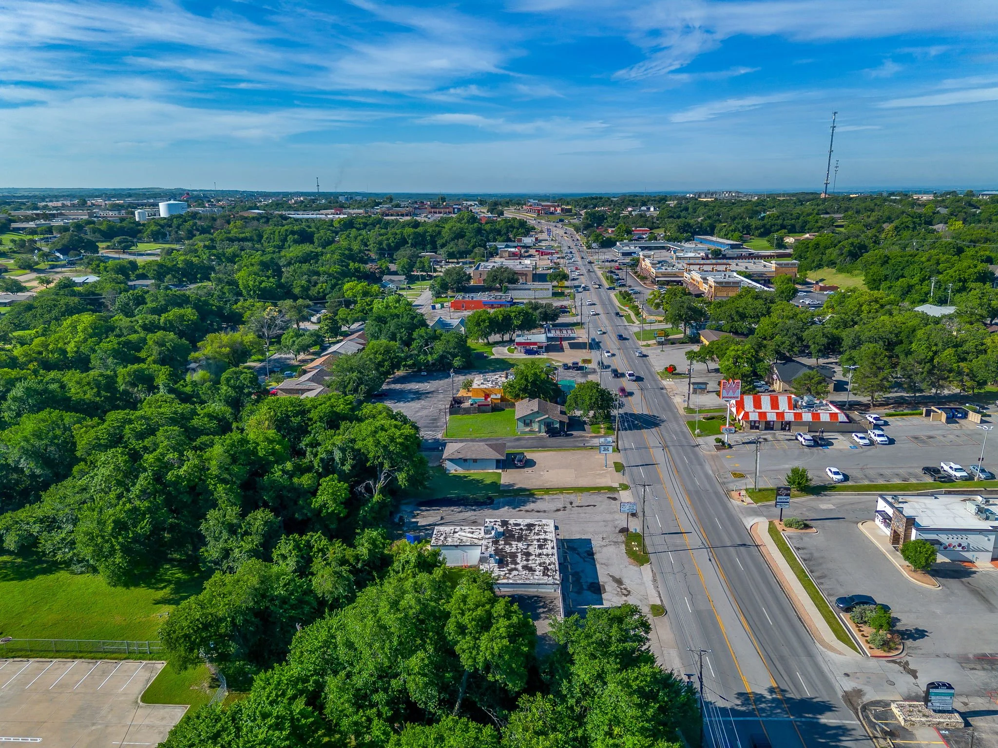 Aerial view of a small town with a main road, surrounded by green trees and residential neighborhoods, under a partly cloudy sky.