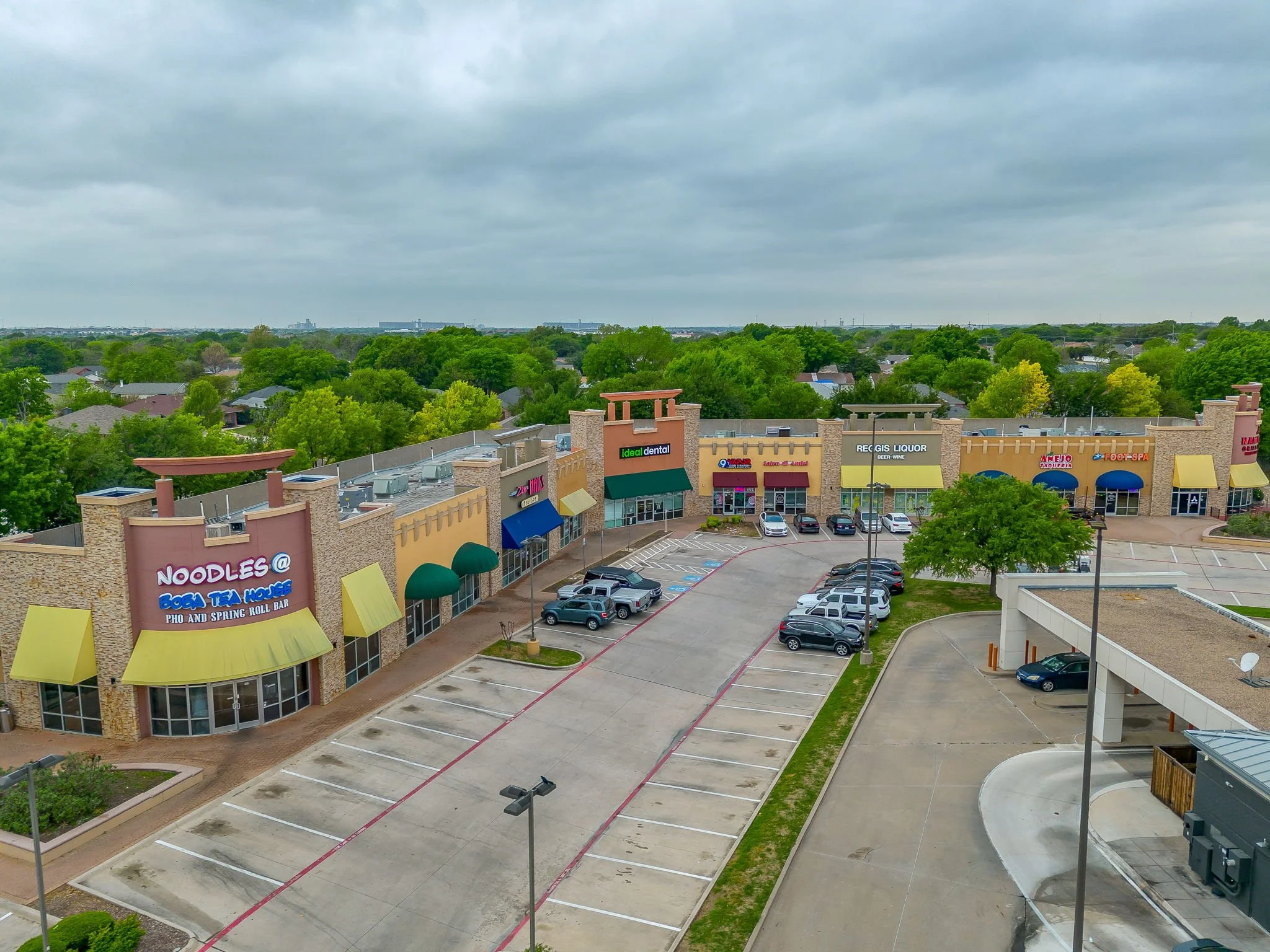 A parking lot in front of shopping stores with no cars parked, surrounded by green trees under a cloudy sky.