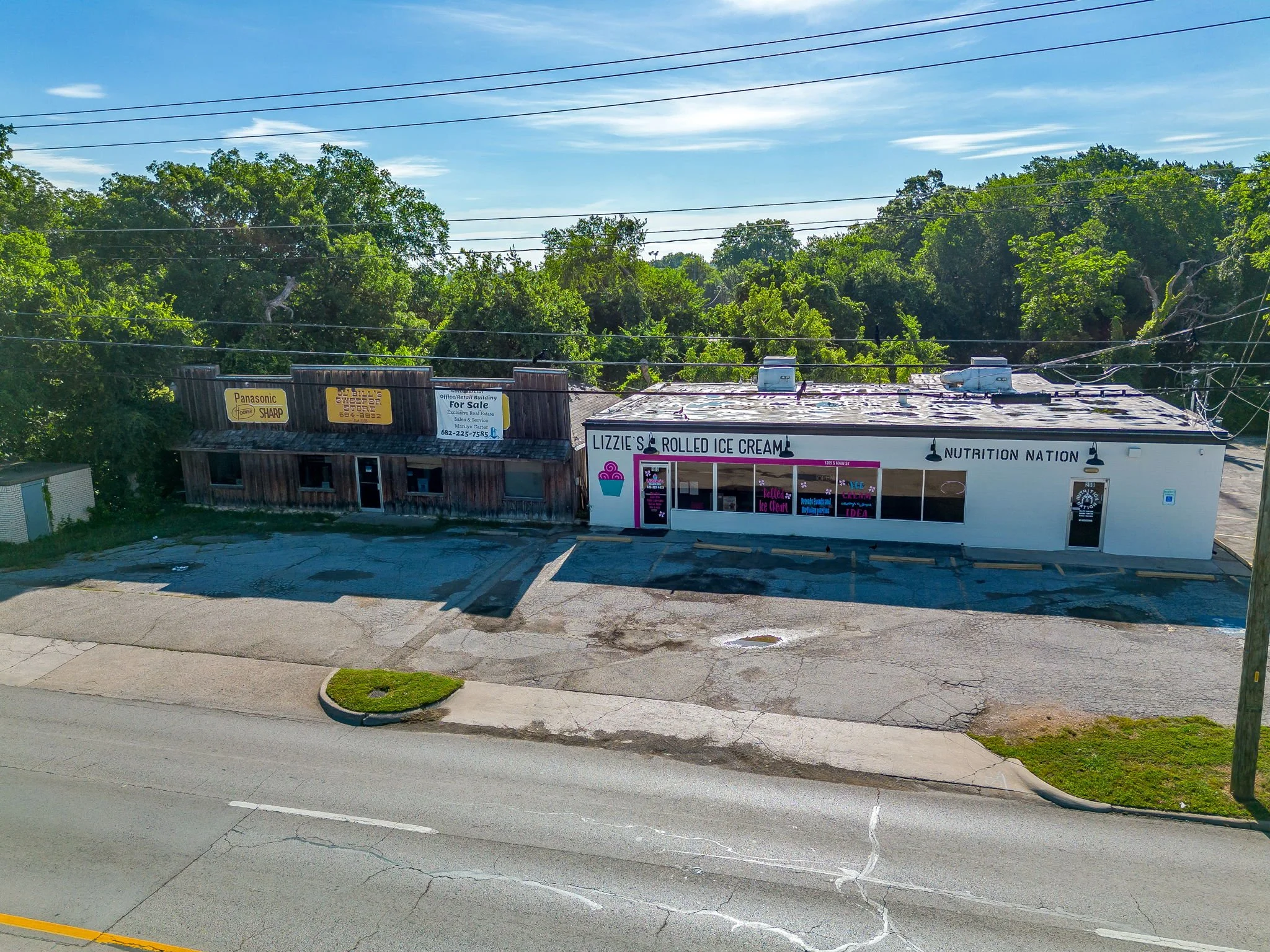 A small commercial building with signage for Lemonade's Rolled Ice Cream, Nutrition Nation, Washington Realty Holding, Office Rental Leasing, and Panasonic Sharp. The building is adjacent to a parking lot and surrounded by green trees, with overhead power lines and a bright blue sky above.