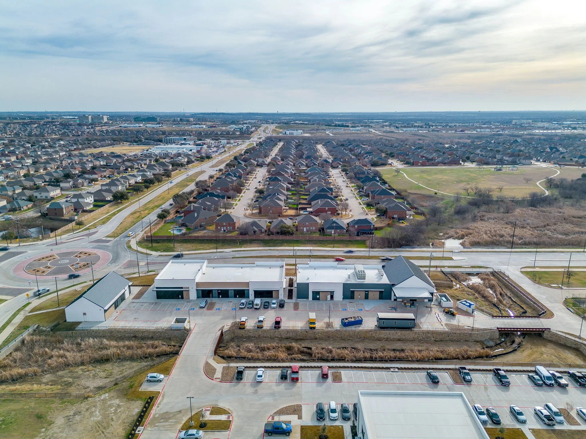 An aerial view of a suburban neighborhood with rows of houses, a parking lot filled with cars, and a park with walking paths and trees.