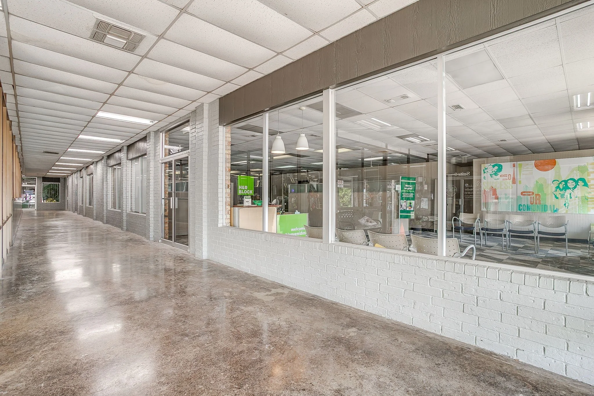 Empty indoor shopping mall corridor with white brick walls, glass storefronts, chairs, and signs inside the stores.