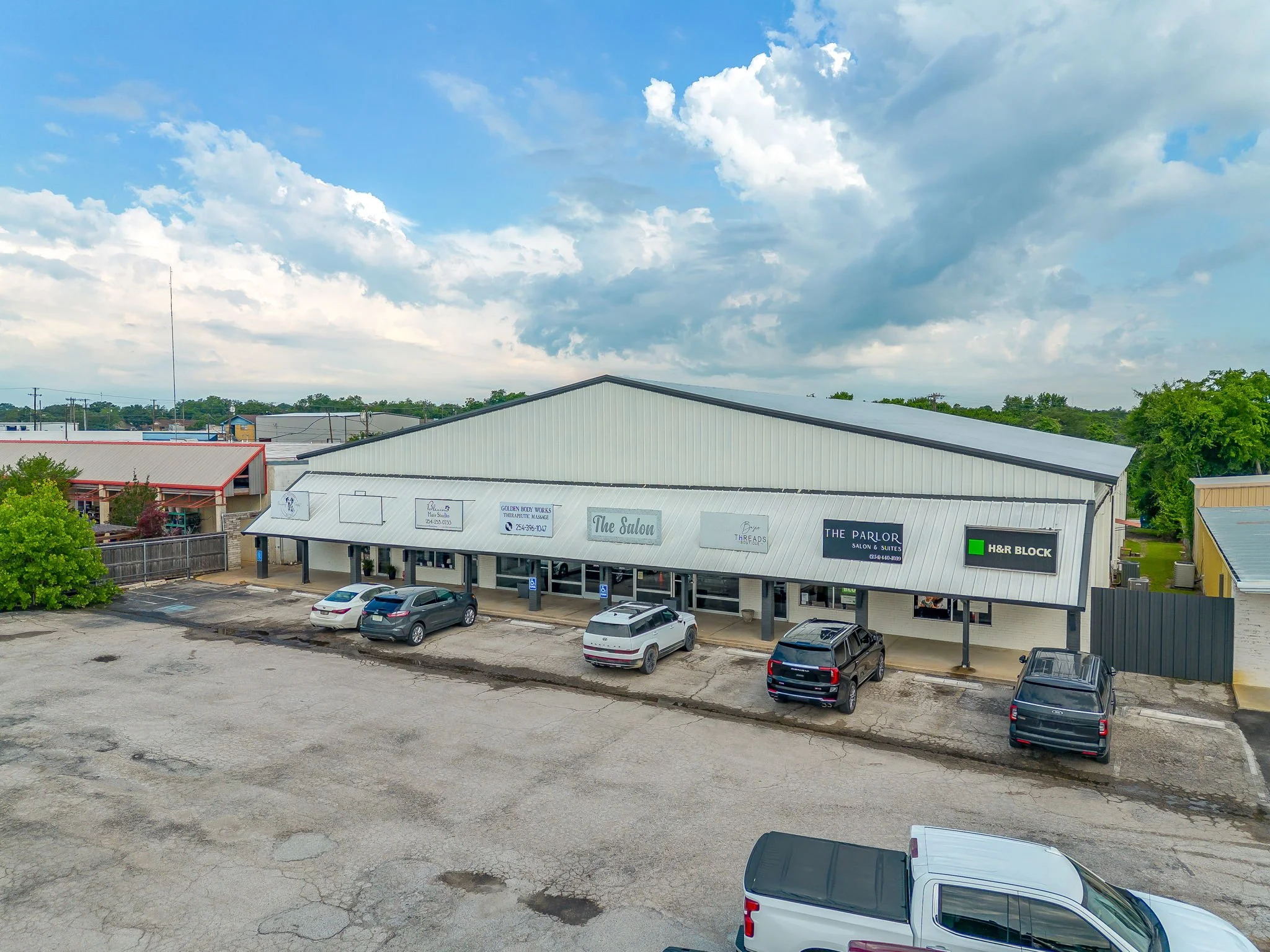 Parking lot with several parked cars in front of a commercial building with a metal roof, under a partly cloudy sky, with some trees and other buildings in the background.