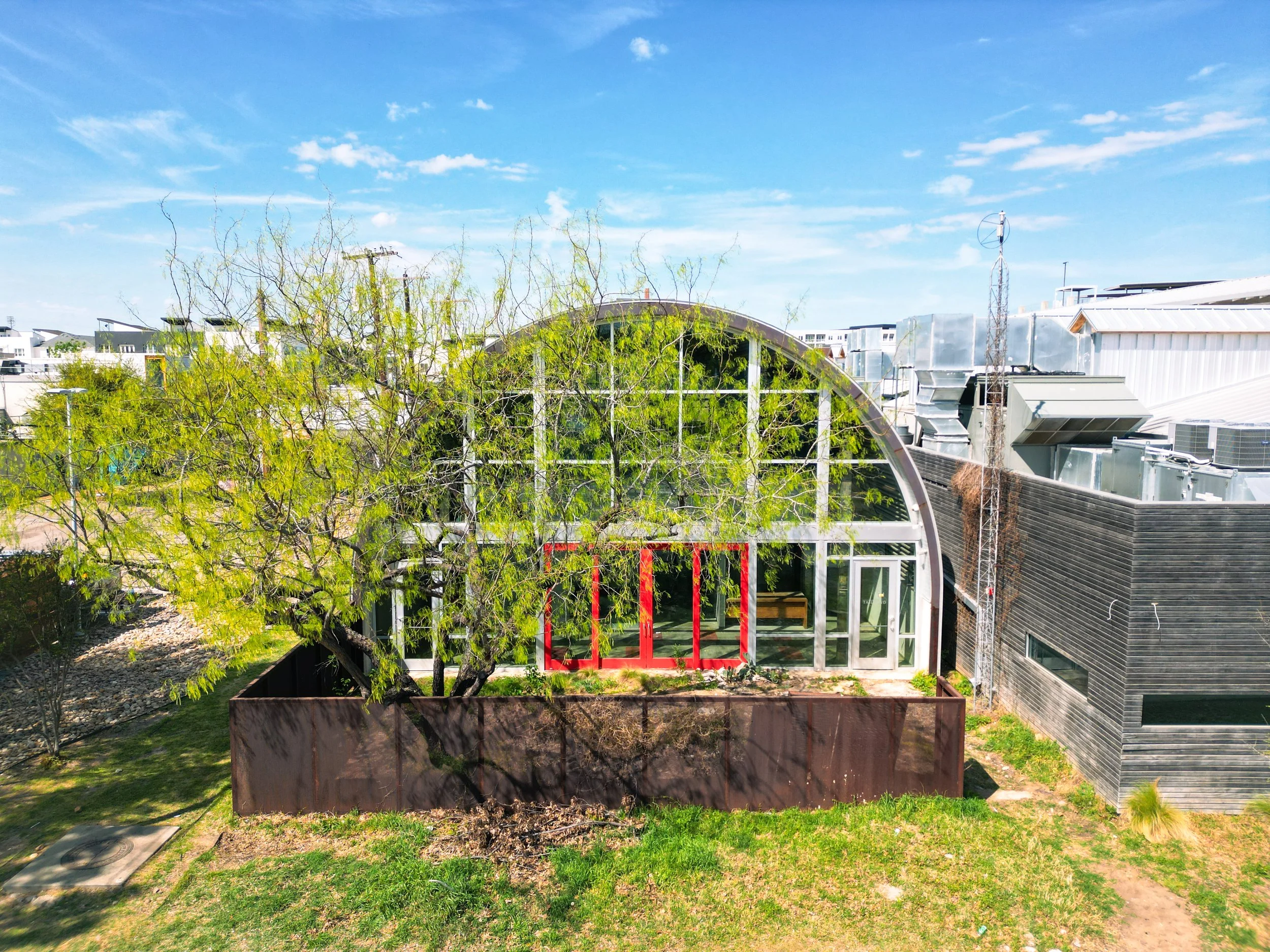 A rooftop view with a modern greenhouse under construction, an arched roof, trees, and buildings, set against a clear blue sky with scattered clouds.