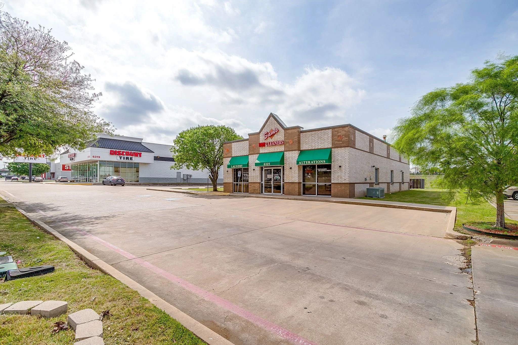 A small shopping plaza with a tire store, a dry cleaners, and a laundromat, surrounded by trees and parking lot on a partly cloudy day.