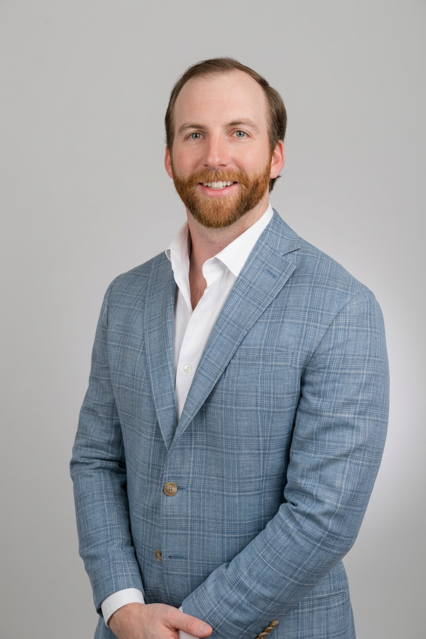 Close-up of a smiling man with blue eyes, reddish-brown beard, wearing a blue checkered blazer and white shirt, crossing arms against a light gray background.