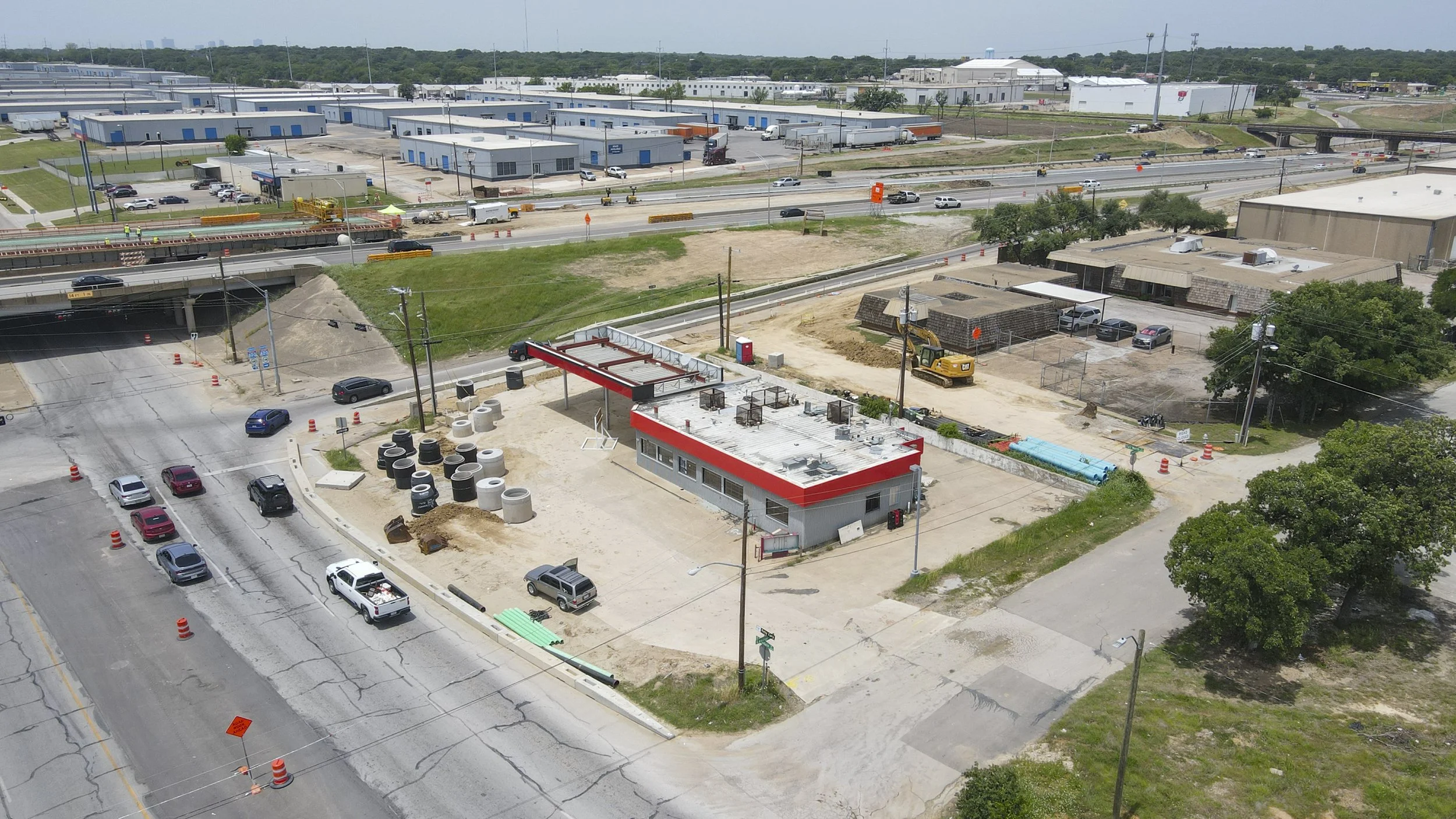 An aerial view of a highway construction site with traffic, construction workers, heavy machinery, and buildings in the background.