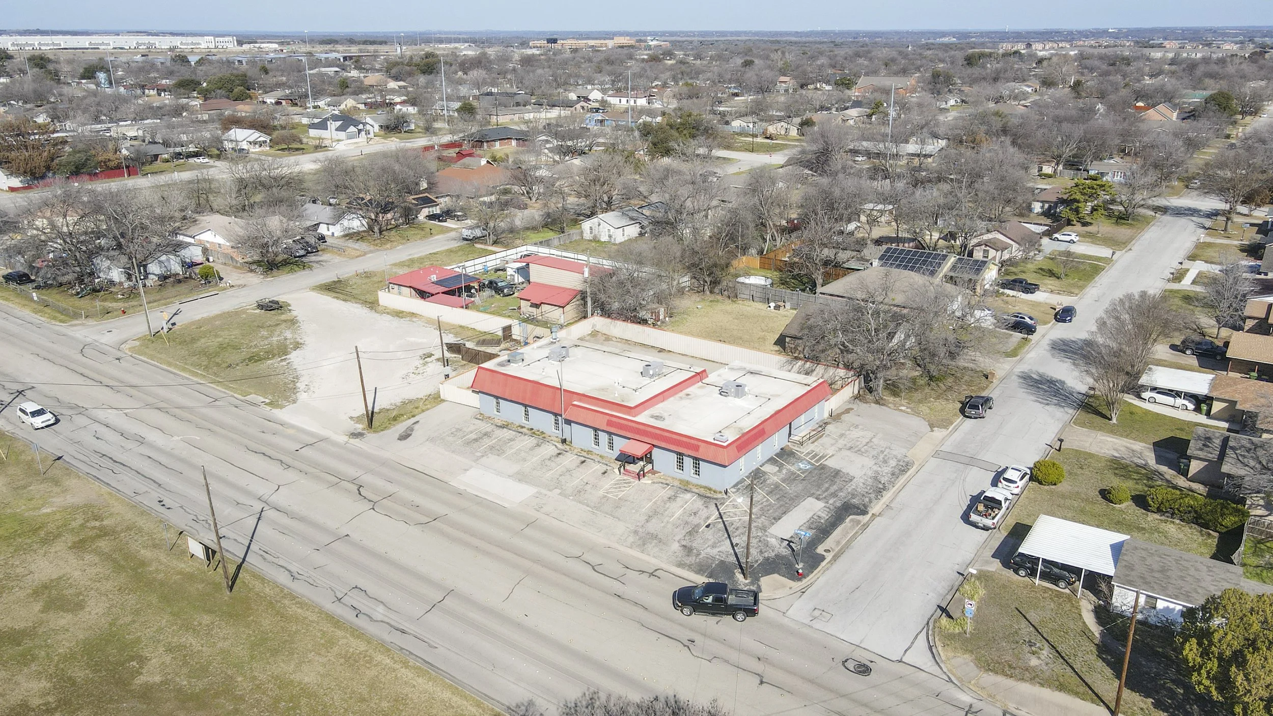 Aerial view of a suburban neighborhood with a mix of houses, trees, and streets. There's a commercial building with a red roof and parking lot in the foreground, and residential homes with various roof colors in the background.