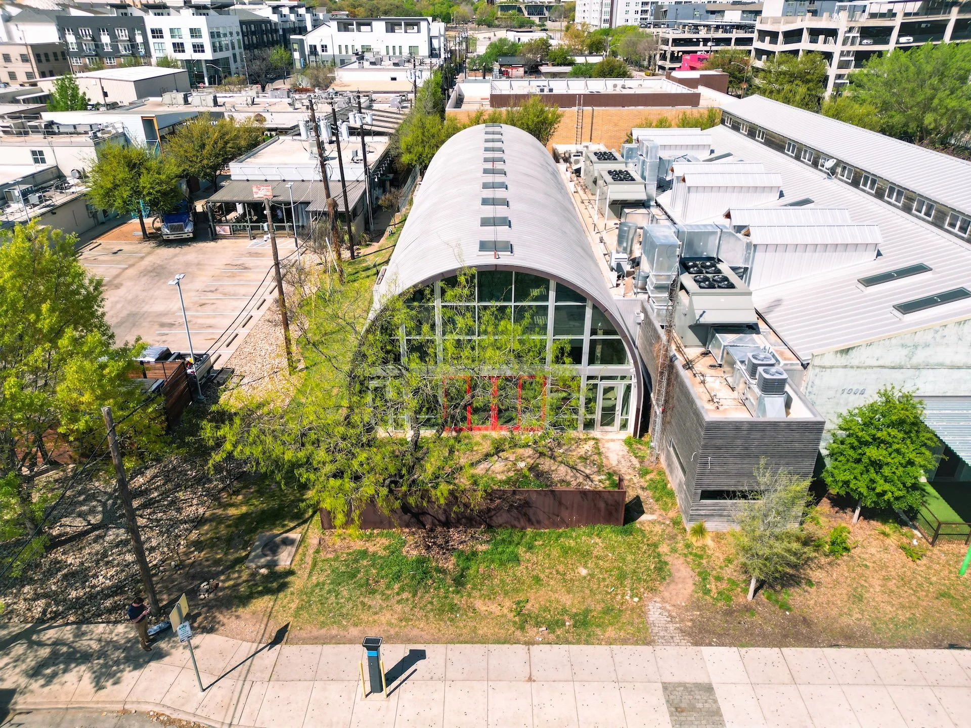 Aerial view of an urban area with modern buildings, green trees, a park, a parking lot, and a large greenhouse with a curved roof and glass exterior.