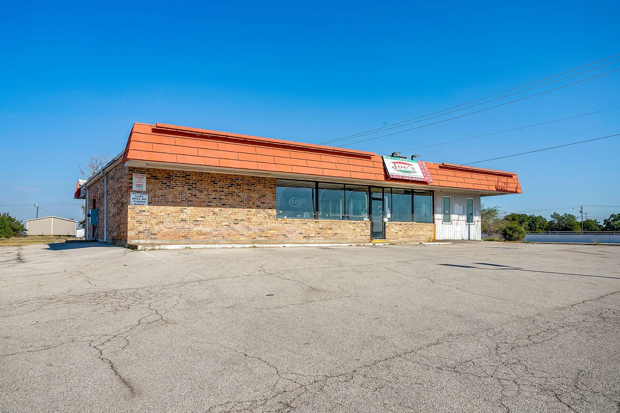 A small brick building with a red metal roof, large windows, and a sign for Joe's Lounge, with a parking lot in front and a clear blue sky overhead.