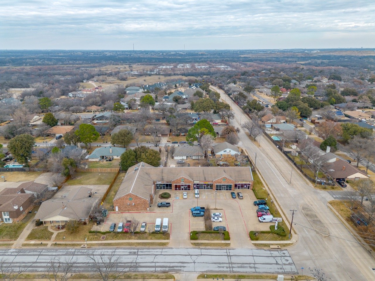 Aerial view of a suburban neighborhood featuring a commercial building with a parking lot, surrounded by residential houses, with trees and streets extending into the distance under a cloudy sky.