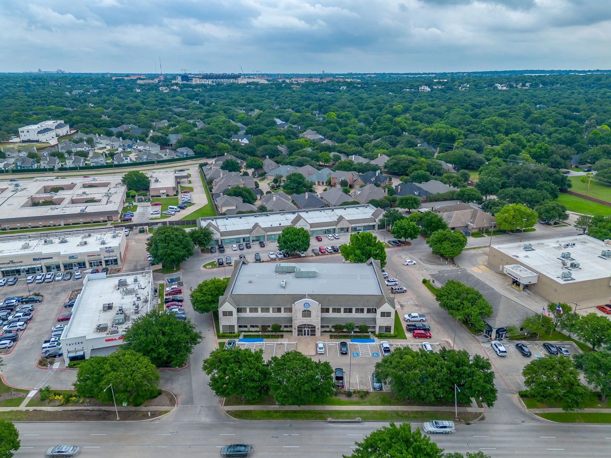 Aerial view of a roadside shopping center with parking lots surrounded by trees and residential houses, with a large green area and a stadium in the distance under a cloudy sky.
