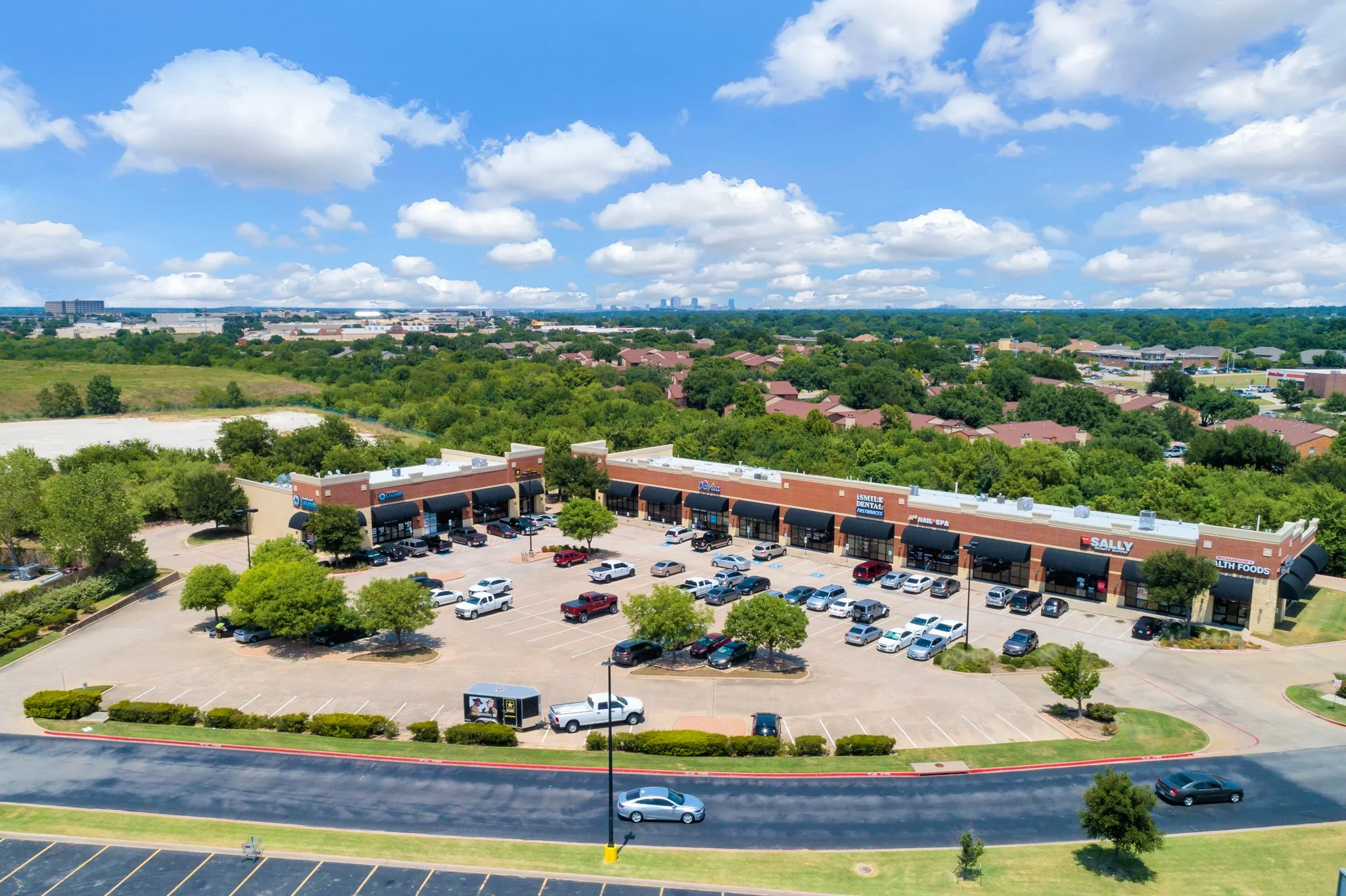 Overhead view of a shopping center parking lot, retail stores, trees, a green area, residential neighborhood, and a distant city skyline under a partly cloudy sky.
