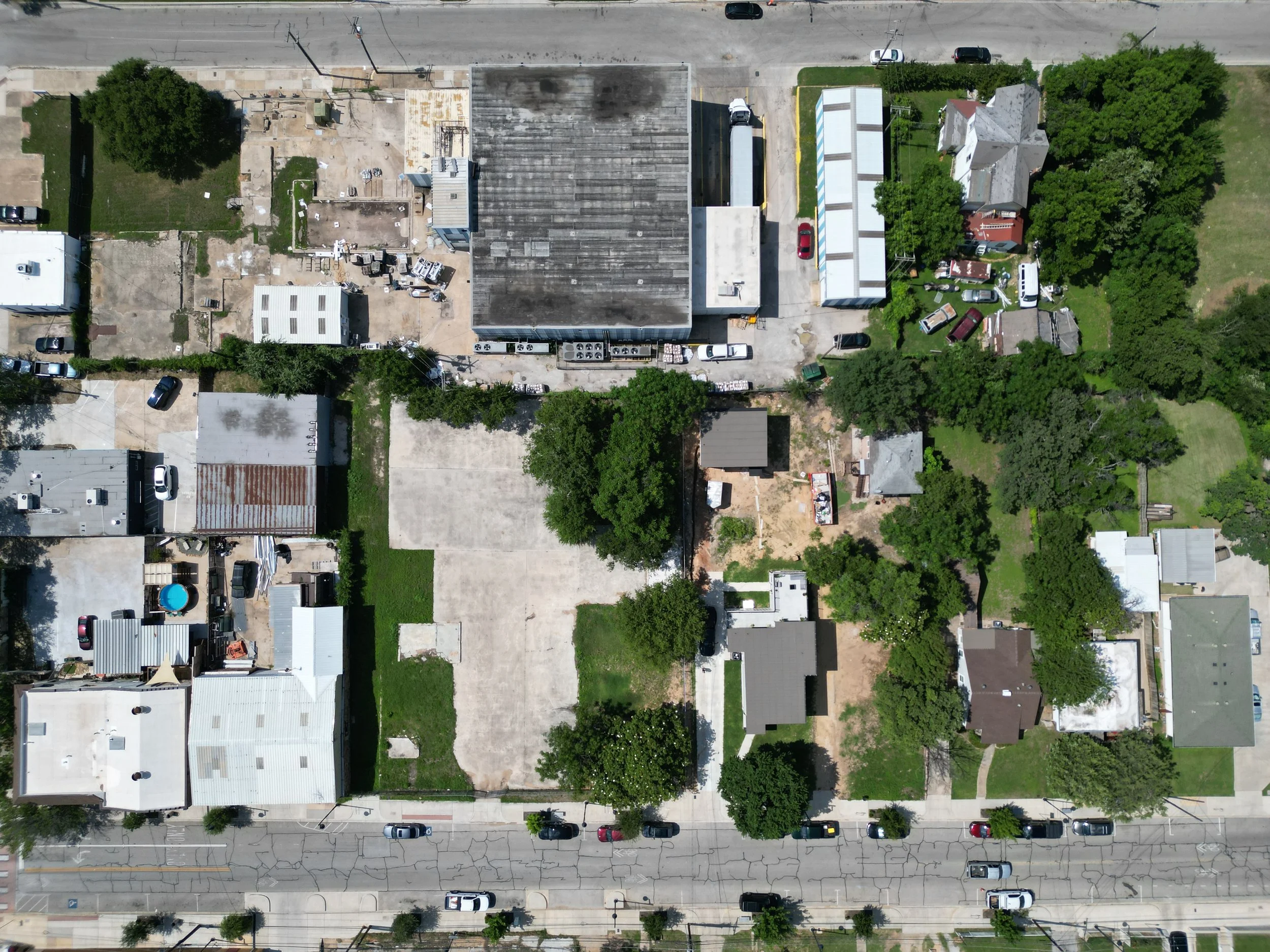 An aerial view of a neighborhood showing various residential buildings, trees, green lawns, parked cars, and a building with a large, dark roof.