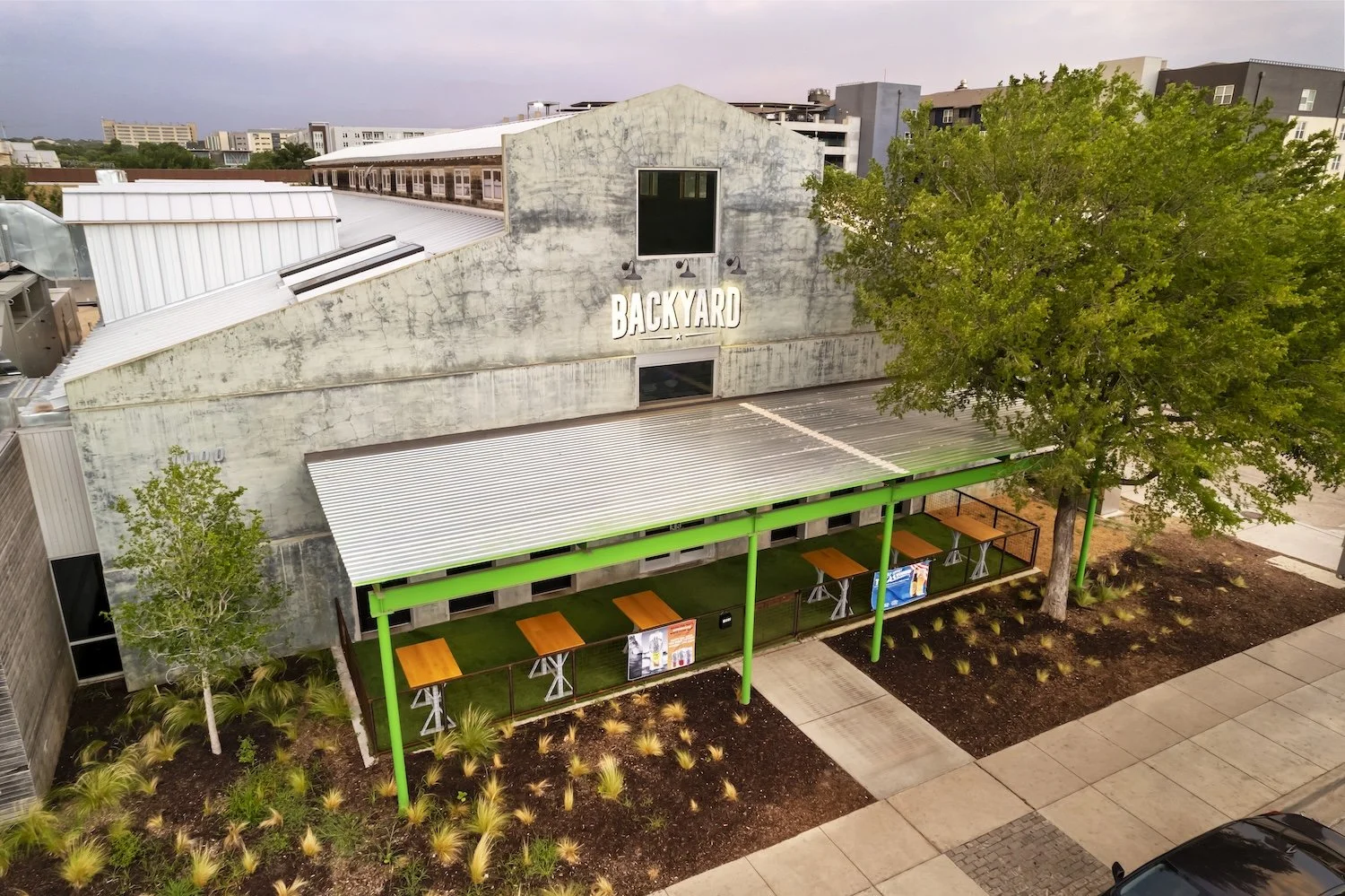 Aerial view of a rustic building with a sign that reads 'BACKYARD', featuring a covered outdoor seating area with wooden tables and green support beams, surrounded by landscaped plants and trees in an urban setting.