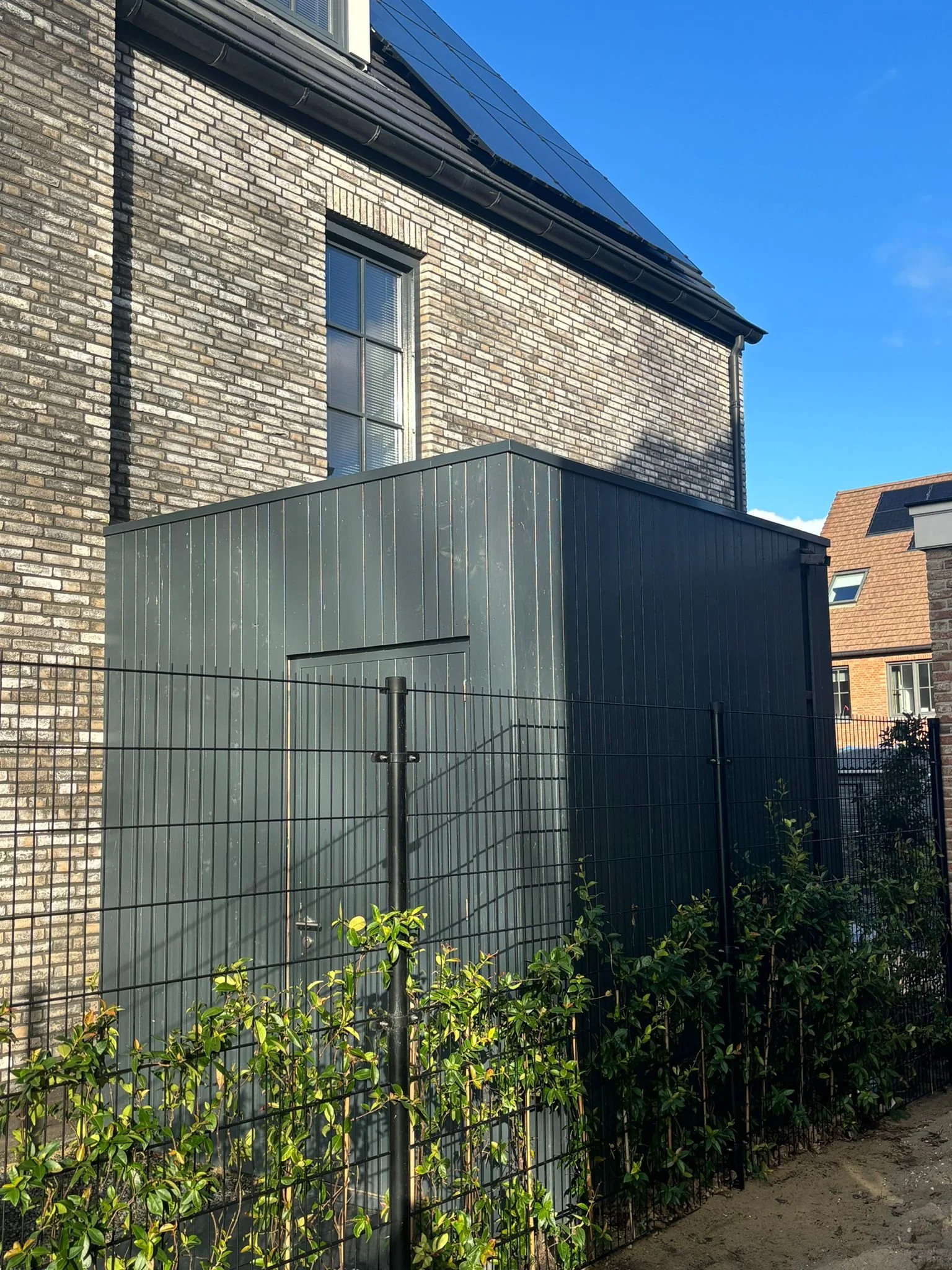 Bouw met groene metalen muur en omheining voor gevel van een huis met bakstenen muur en raam, onder een blauwe hemel.