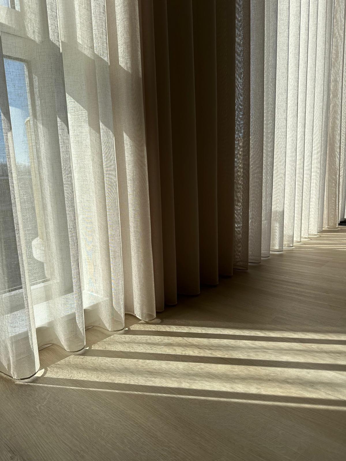 Licht door een raam met witte en beige gordijnen, dat schaduwpatronen op de houten vloer werpt.