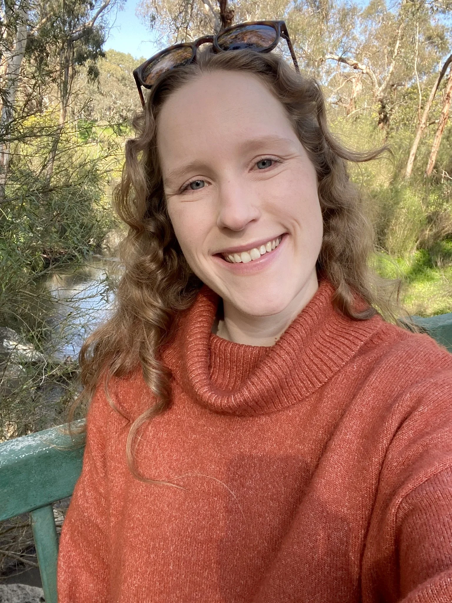 A smiling woman with curly hair, wearing sunglasses on her head and an orange turtleneck sweater, taking a selfie outdoors near a small creek surrounded by trees.