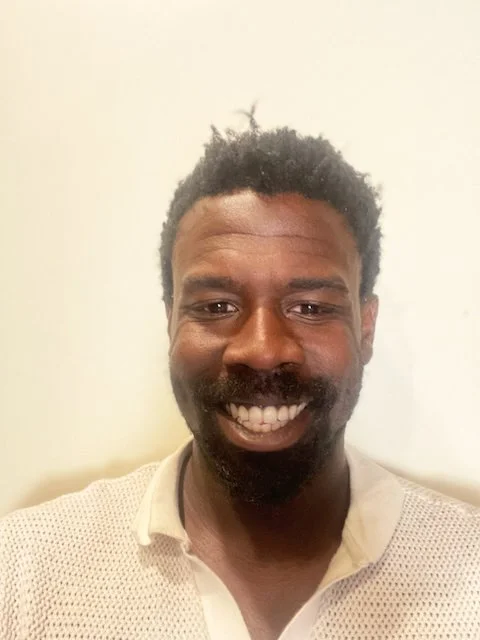 A smiling Black man with short curly hair and a beard, wearing a beige button-up shirt, standing against a plain white wall.