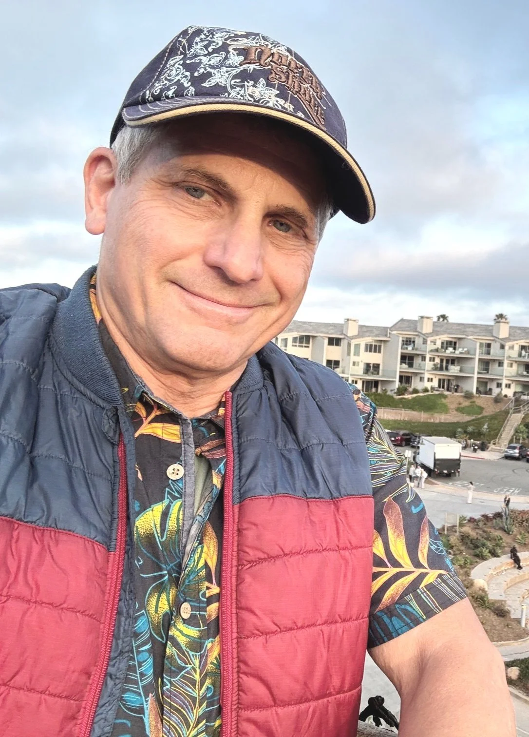 A man smiling outdoors, wearing a colorful hat and a red and black jacket, with a residential area in the background.
