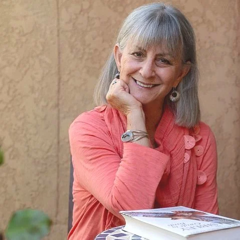 Smiling woman with gray hair wearing a coral blouse, resting her chin on her hand, sitting at a table with a book in front of her.