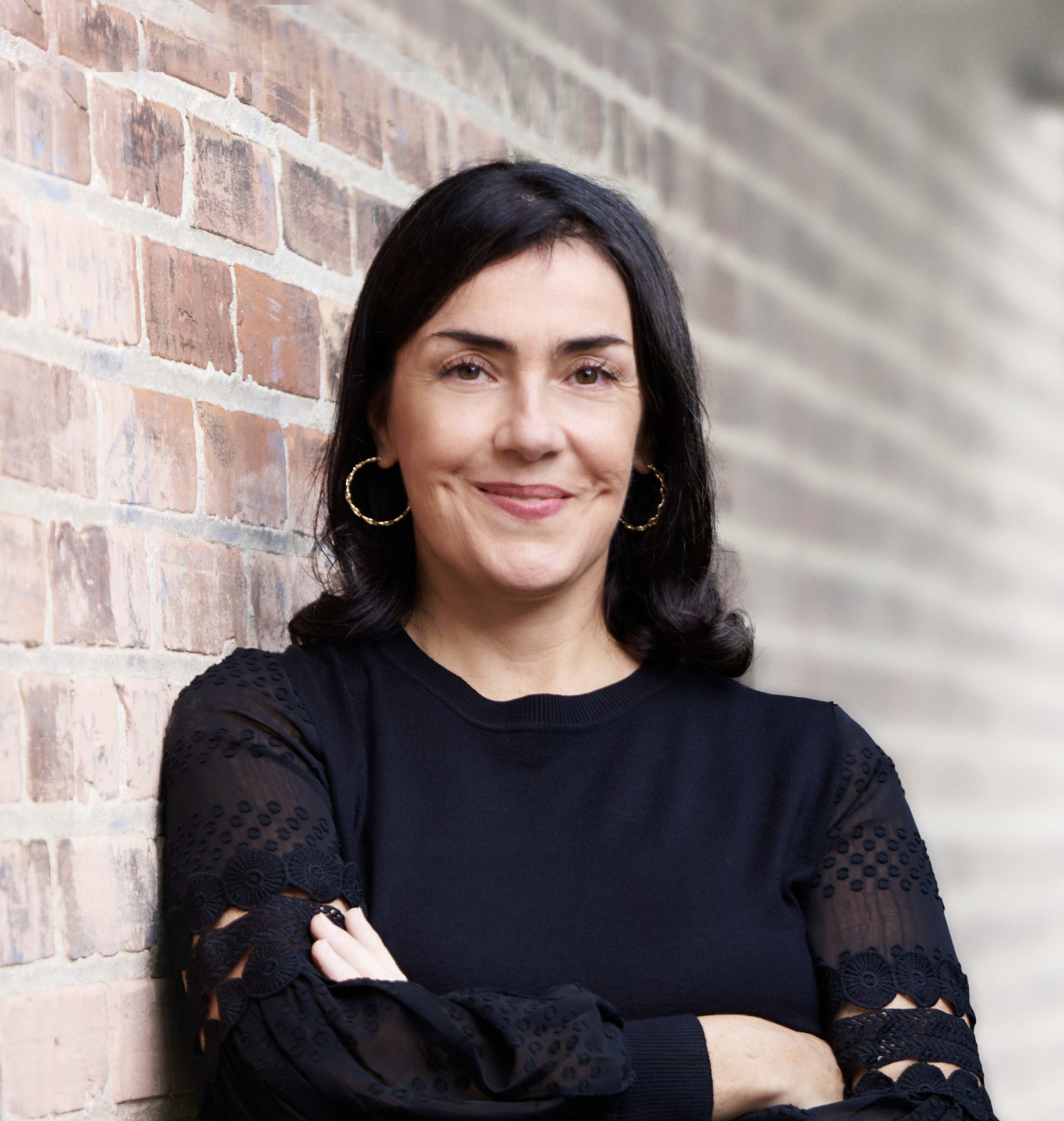 A woman with dark hair wearing a black top and hoop earrings, standing against a brick wall with her arms crossed and smiling at the camera.