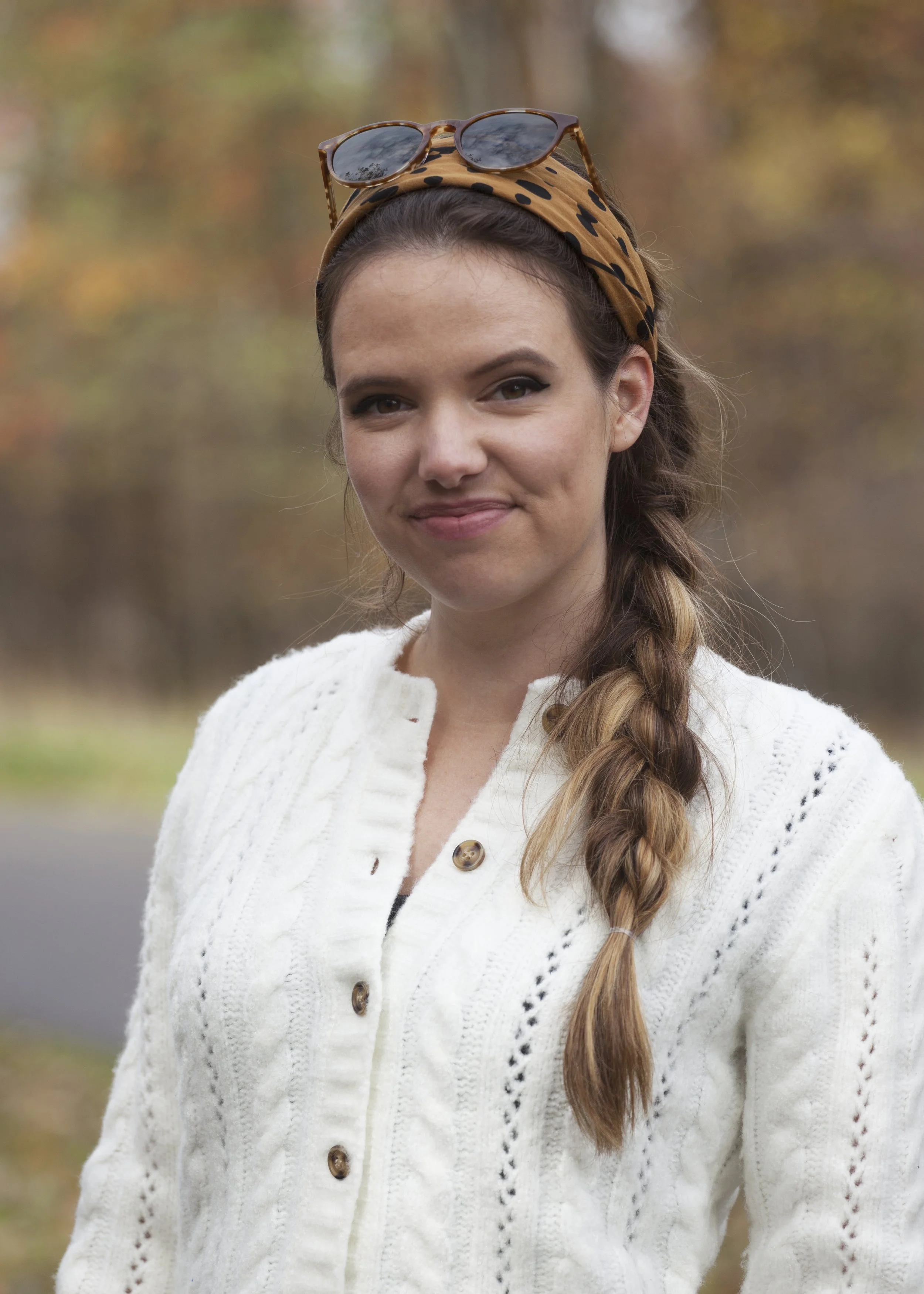 A young woman with long braided hair wearing a white knit sweater, a leopard print headband with sunglasses on top, standing outdoors with blurred autumn trees in the background.