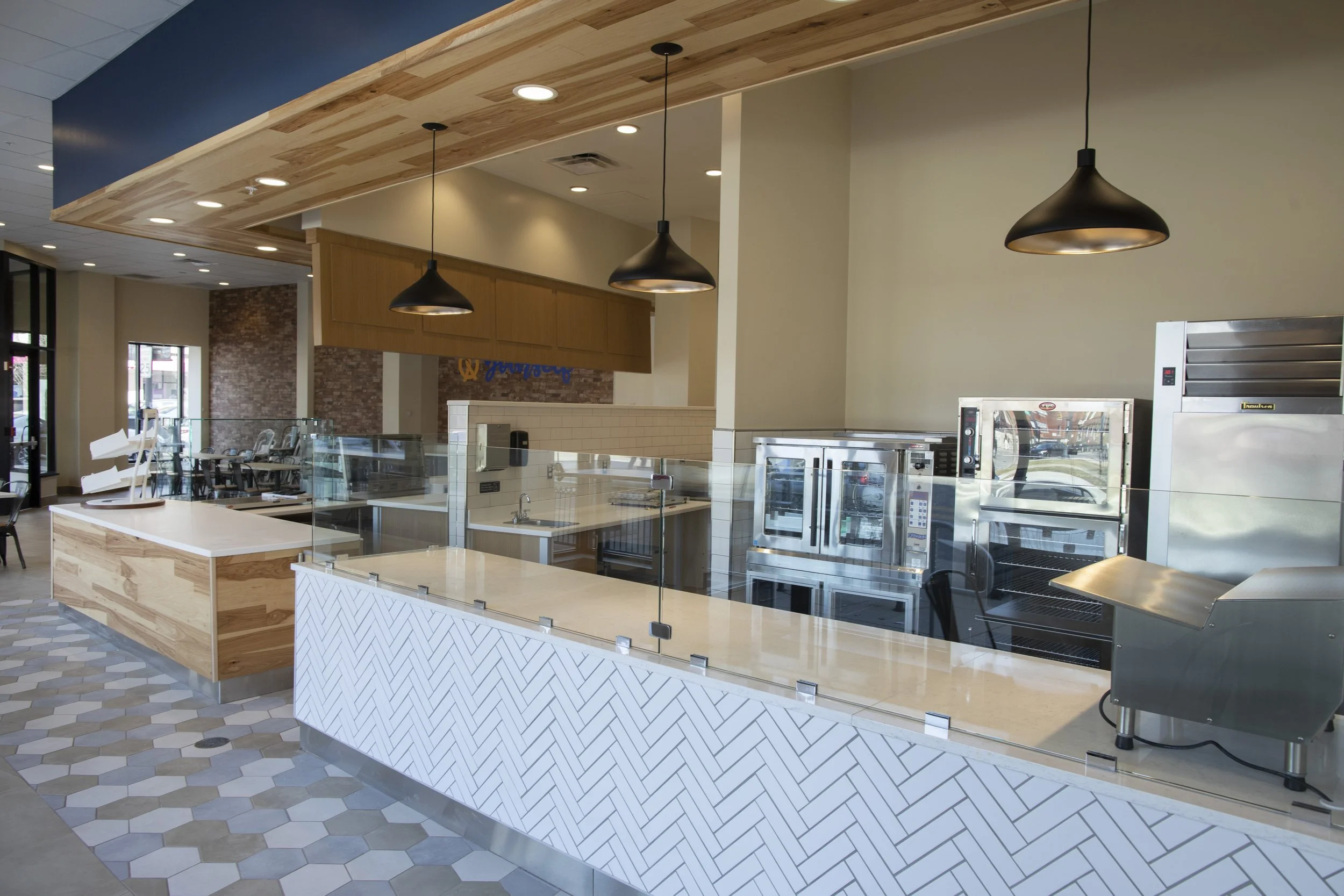 Interior of a modern commercial kitchen with glass counter, ovens, and stainless steel appliances, featuring wood accents and pendant lights.