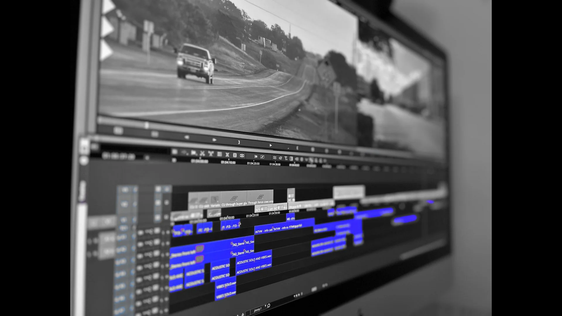Close-up of a computer screen displaying a video editing timeline with multiple blue and gray clips, and a black and white landscape of a curvy road with a car in the distance.