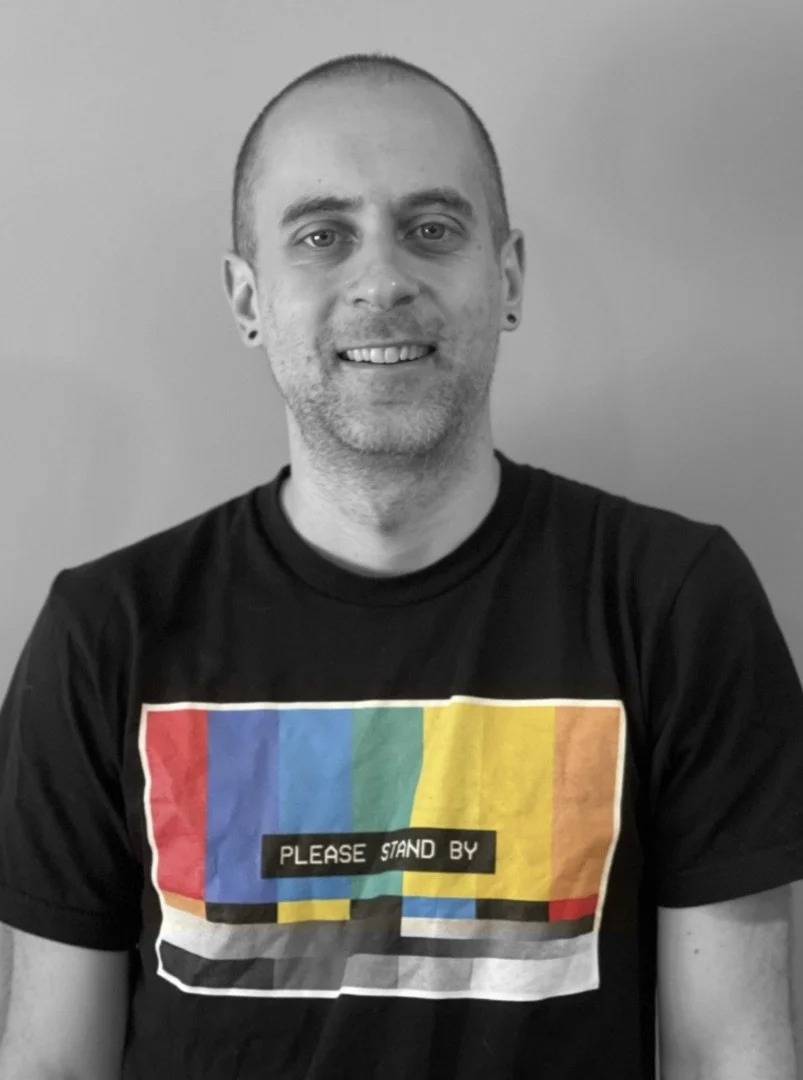 Black and white photo of a man with short hair, wearing a T-shirt with a colorful static television screen graphic and the words "Please Stand By" on it, smiling at the camera.