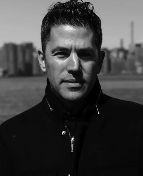 Black and white photo of a man with short dark hair, wearing a jacket, outdoors with city skyline in the background.