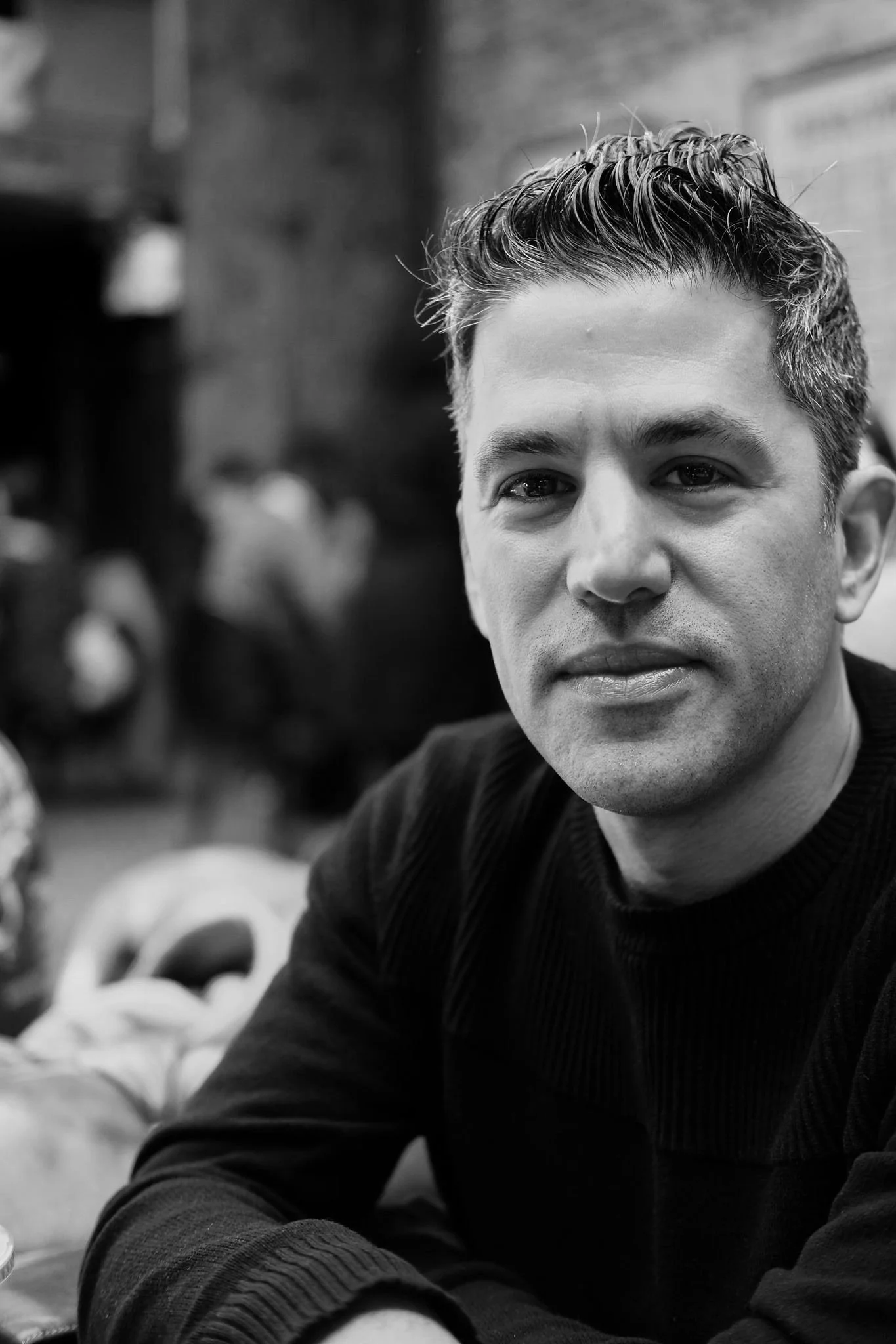 Black and white close-up portrait of a man with short, styled hair, wearing a dark sweater, looking directly at the camera with a neutral expression. Background features blurred figures in an indoor setting.