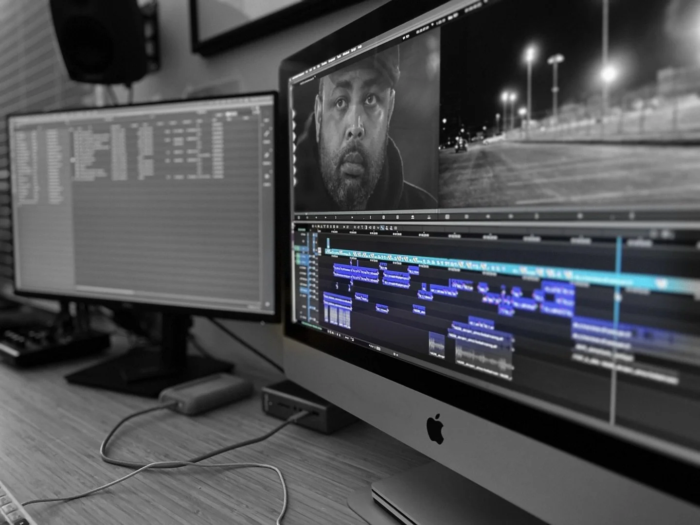 A computer workspace with two monitors; the right monitor displays video editing software with a man’s face and a nighttime street scene, while the left monitor shows a file browser or media library. The desk has a keyboard, mouse, and cables, indicating a video editing or multimedia editing setup.